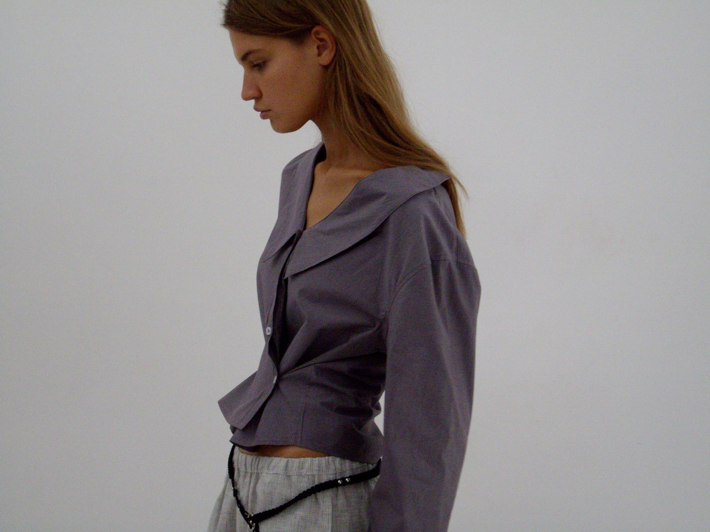 A woman with long brown hair stands in profile against a plain background, wearing the Waist Button Shirt - Currant by Deiji Studios and light gray pants, with a black beaded accessory around her waist.