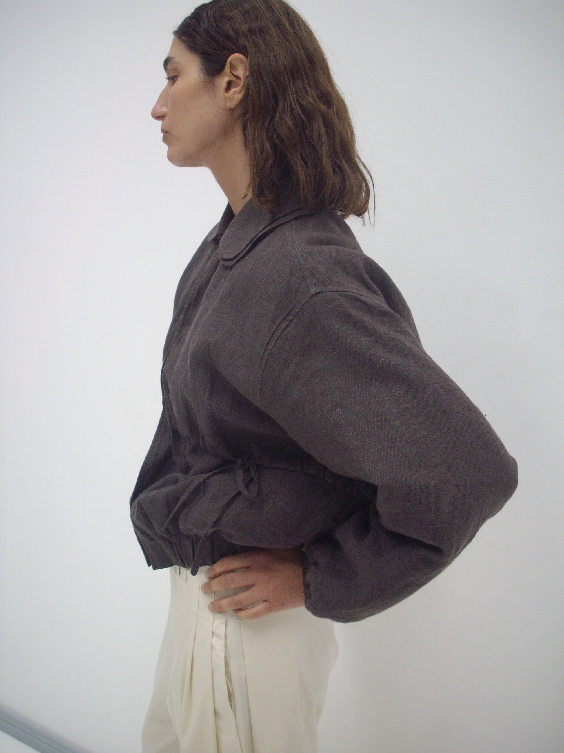 A person with wavy brown hair, wearing a loose Deiji Studios Double Layered Jacket - Bark and cream pants, stands in profile against a plain white background.