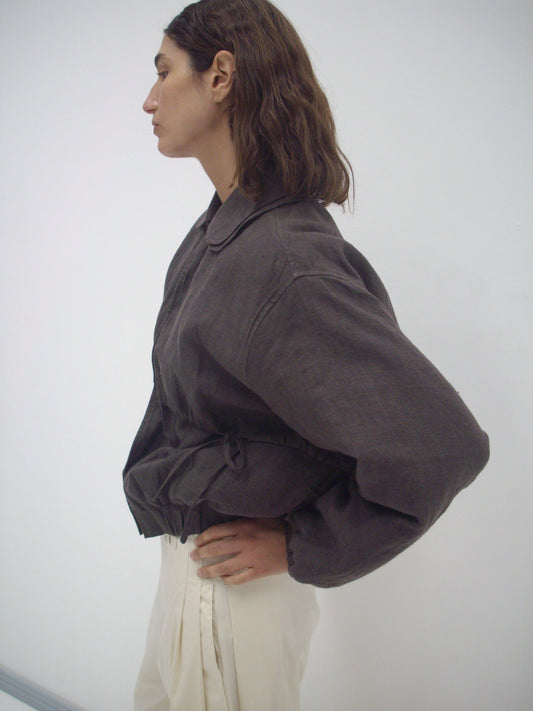 A person with wavy brown hair, wearing a loose Deiji Studios Double Layered Jacket - Bark and cream pants, stands in profile against a plain white background.
