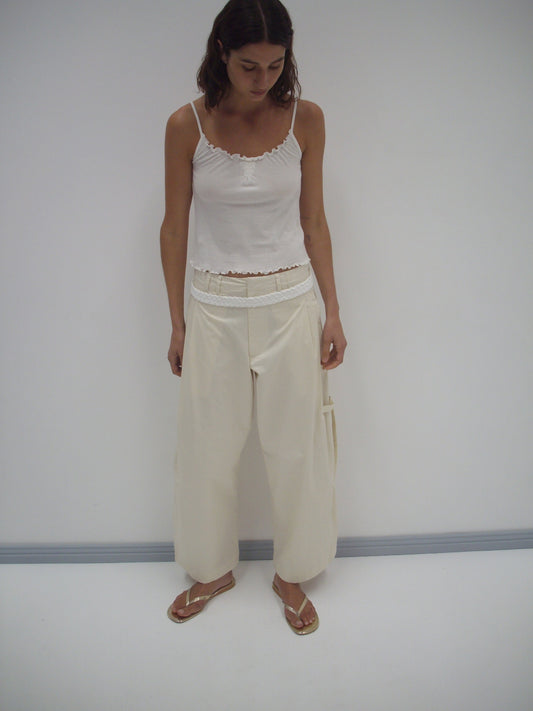 A woman stands indoors against a plain white wall, wearing Deiji Studios’ Tab Pant in off-white, paired with a white camisole and gold sandals. She looks down with her hands by her sides.