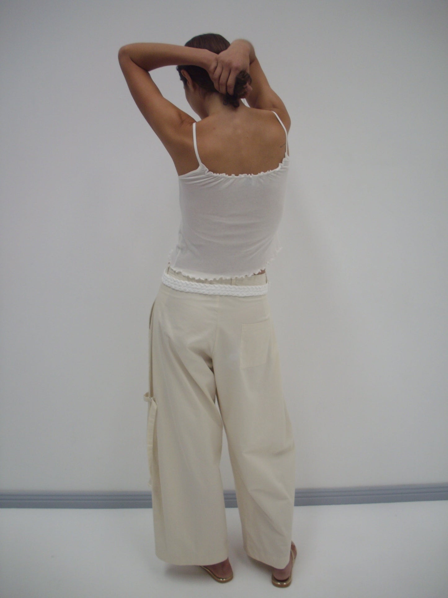 A person in a white tank top and the Tab Pant - Off White, made from organic cotton, stands with their back to the camera and arms raised against a light wall—an effortless look from Deiji Studios.