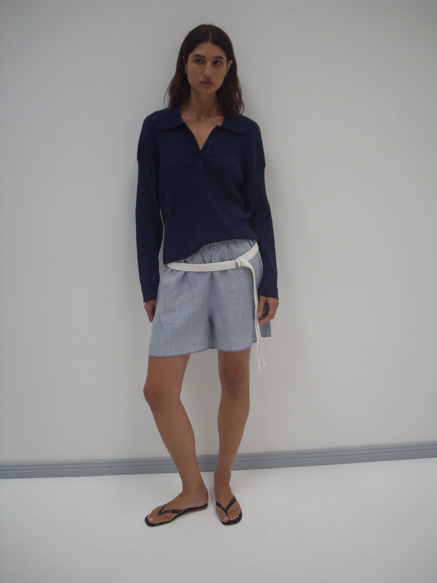 A woman with long brown hair stands against a white wall, wearing the Polo Knit - Dark Blue long-sleeve shirt, light gray shorts with a white belt, and black flip-flops in a relaxed pose.