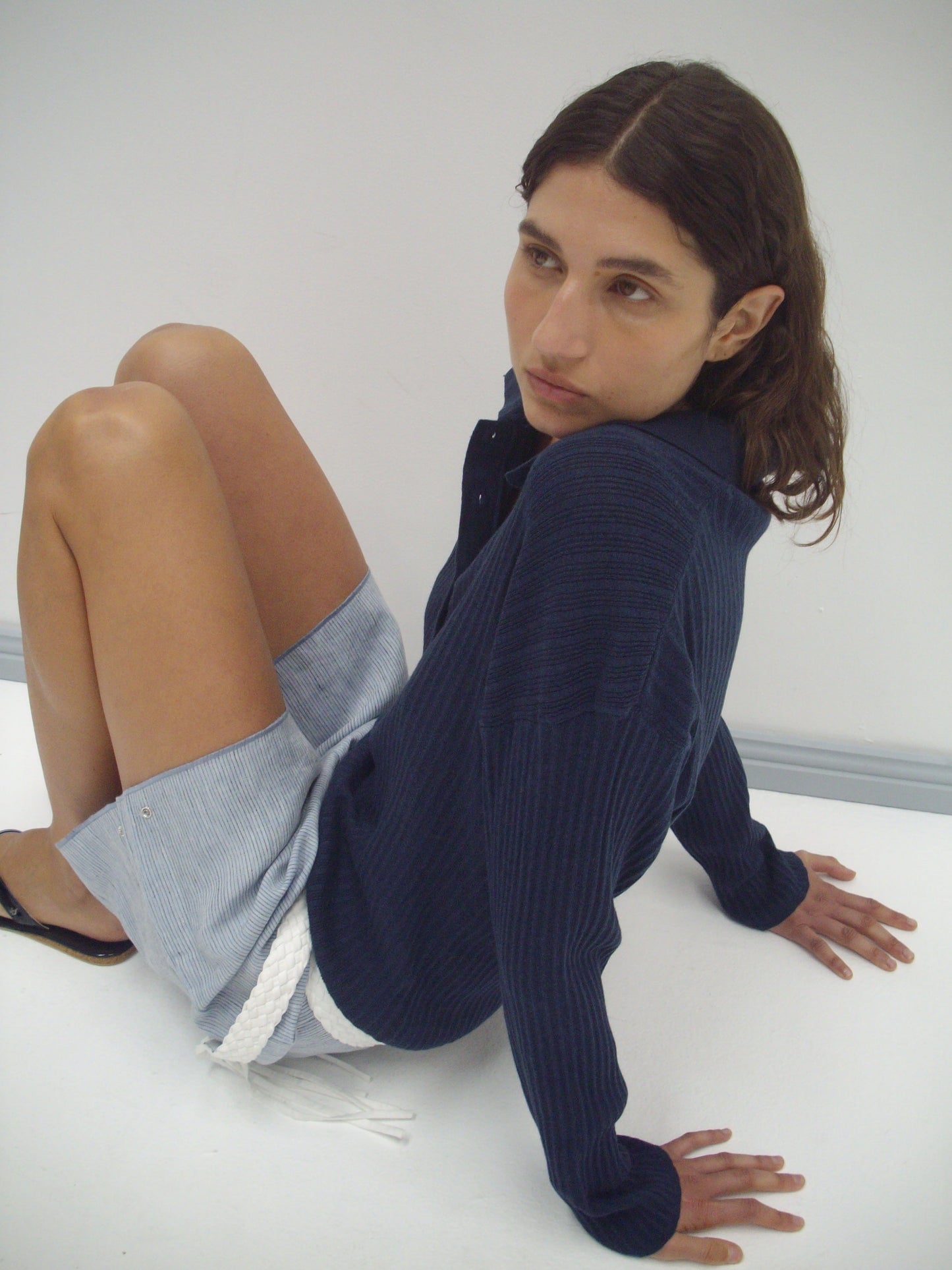 A person with wavy brown hair sits on the floor against a white background, wearing a dark blue Polo Knit from Deiji Studios and light blue shorts with a white rope belt.