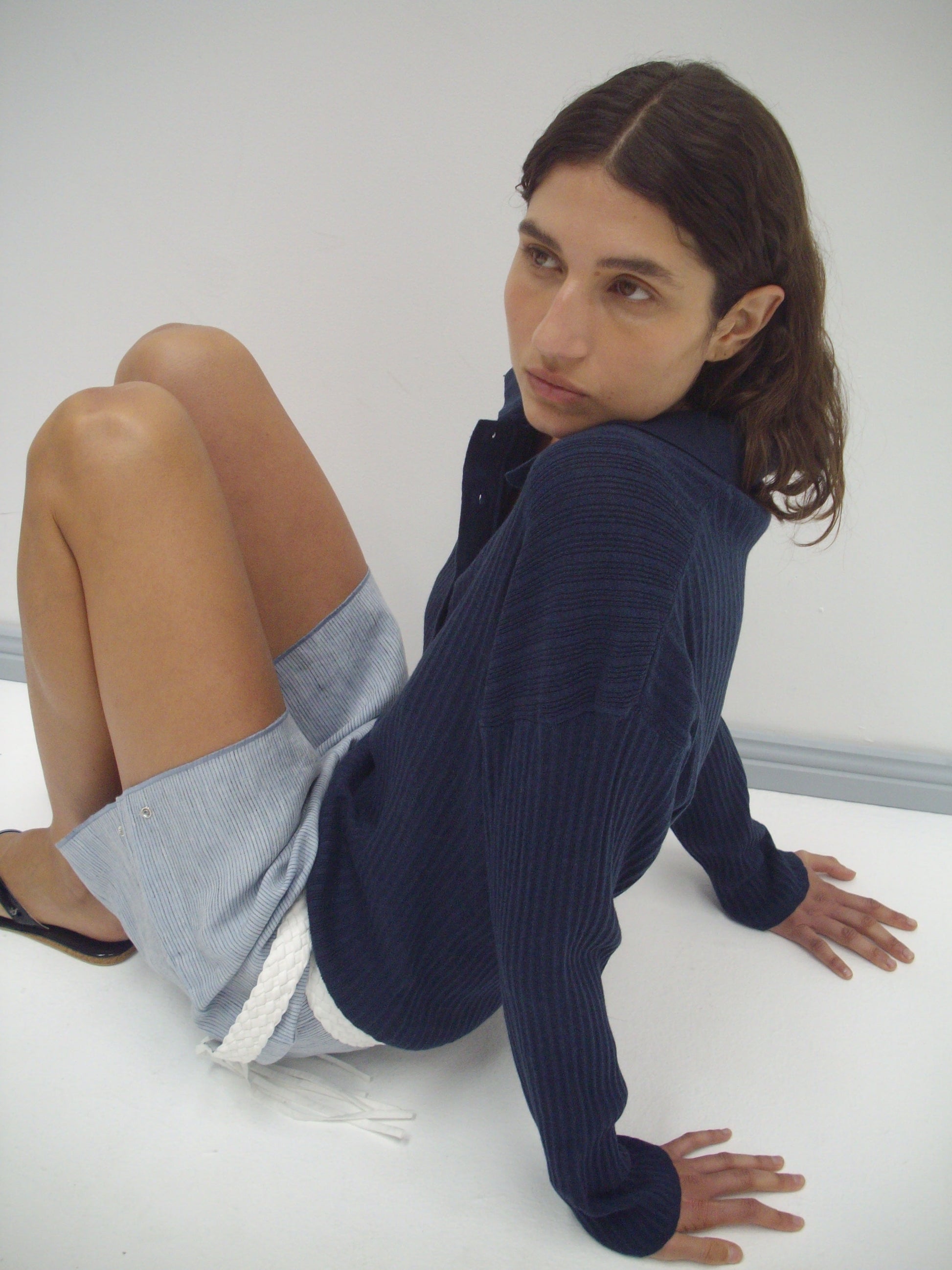 A person with wavy brown hair sits on the floor against a white background, wearing a dark blue Polo Knit from Deiji Studios and light blue shorts with a white rope belt.