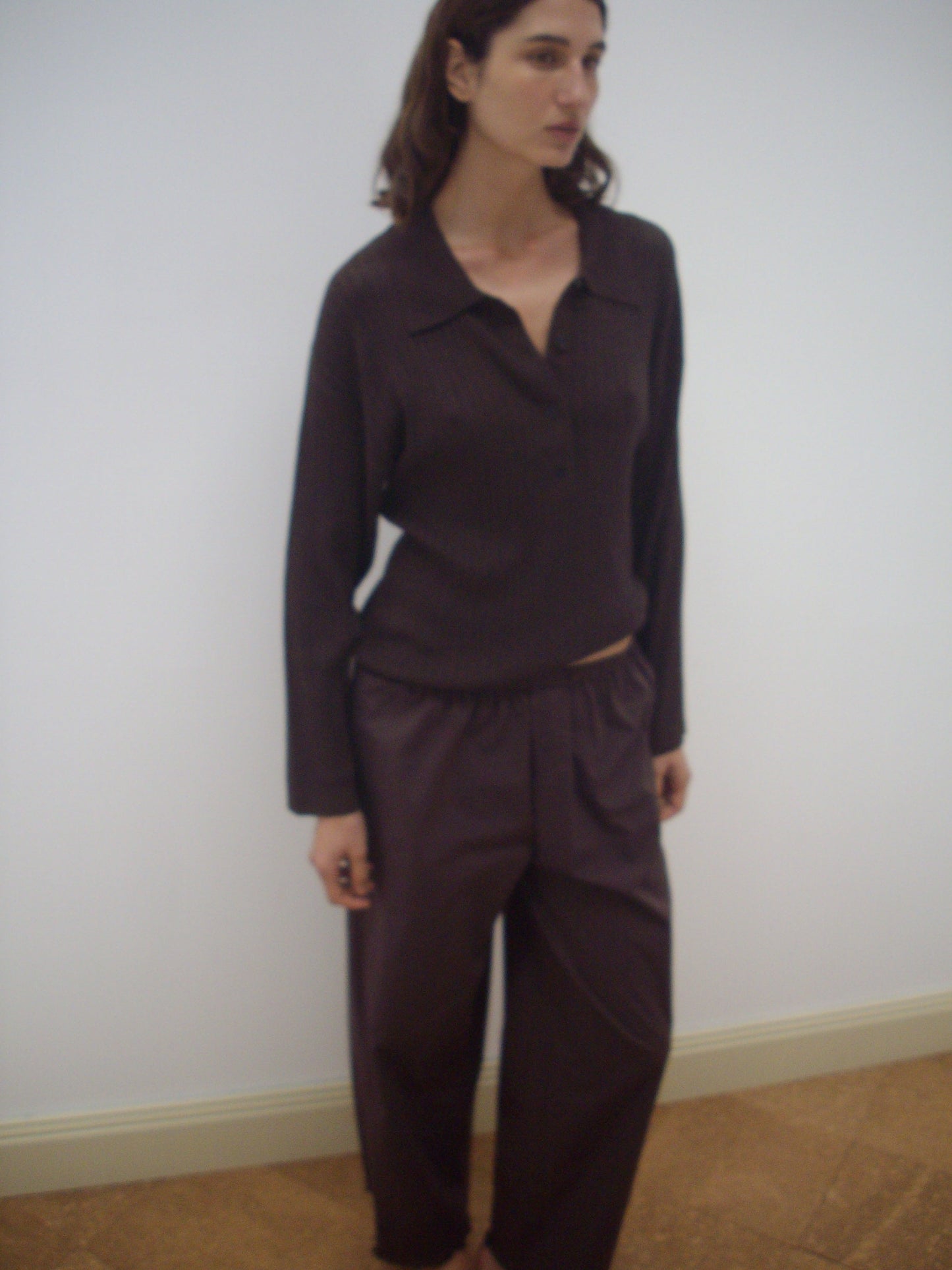 A woman stands indoors against a plain light-colored wall, wearing a loose-fitting Deiji Studios Polo Knit - Dark Almond long-sleeve top and matching wide-leg pants crafted from organic cotton.