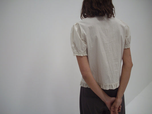 A person with shoulder-length brown hair wears the Pintuck Top - Bone Check and dark pants, standing with their back to the camera and hands clasped behind them against a plain white background.
