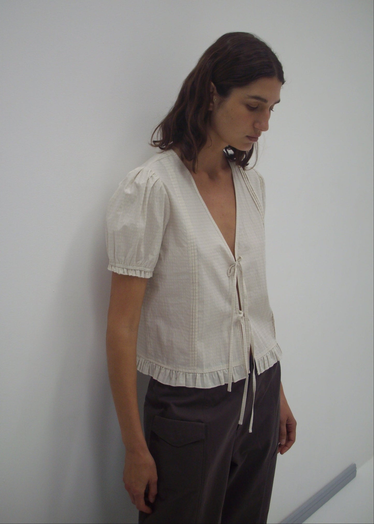 Model wearing the Pintuck Top - Bone Check by Deiji Studios, featuring a cream linen blend with blouson sleeves, ruffled hem, deep V-neck, and tie front detail, paired with dark grey trousers against a white wall.