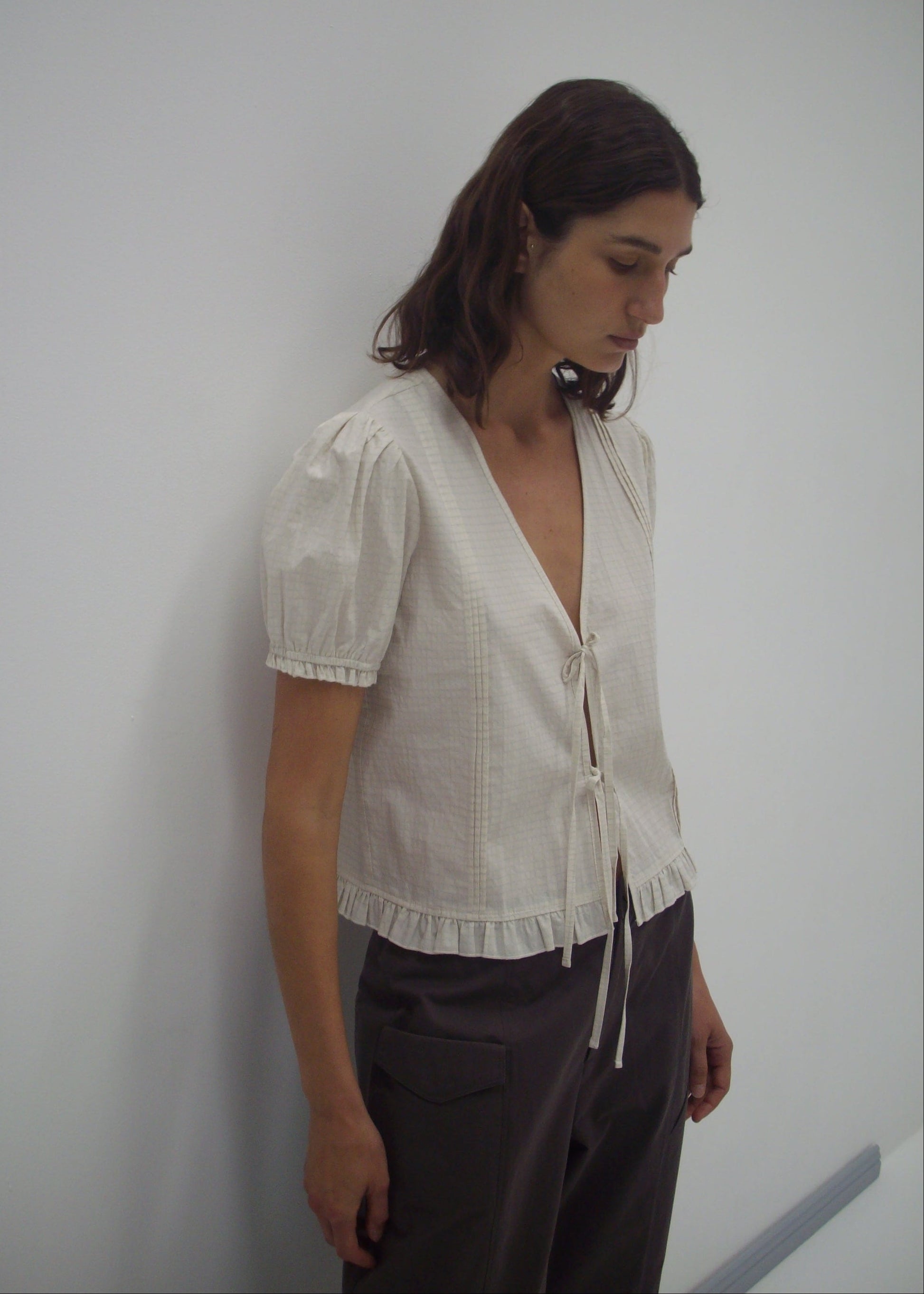 Model wearing the Pintuck Top - Bone Check by Deiji Studios, featuring a cream linen blend with blouson sleeves, ruffled hem, deep V-neck, and tie front detail, paired with dark grey trousers against a white wall.