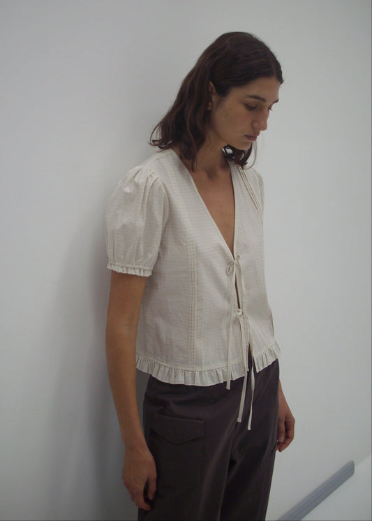Model wearing the Pintuck Top - Bone Check by Deiji Studios, featuring a cream linen blend with blouson sleeves, ruffled hem, deep V-neck, and tie front detail, paired with dark grey trousers against a white wall.