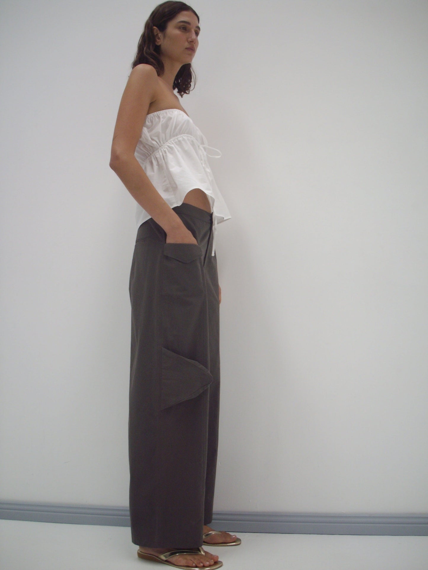 A woman with medium-length dark hair and a neutral expression stands sideways against a white wall, wearing a Deiji Studios strapless cotton top in white, loose dark gray pants with large front pockets, and flat sandals.