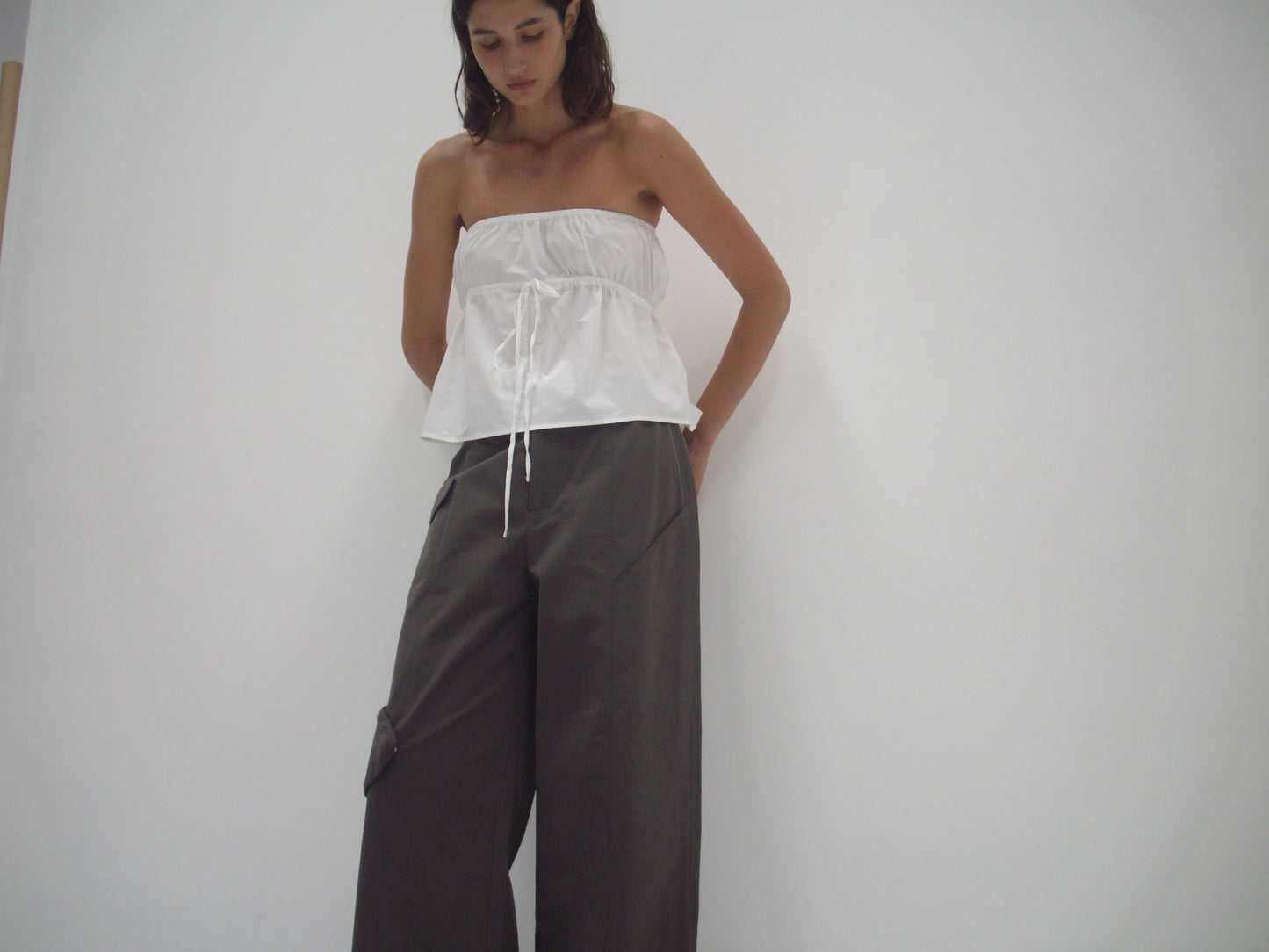 A woman stands against a white wall, looking down. She wears the Deiji Studios strapless cotton top - white and loose-fitting dark gray cargo pants.