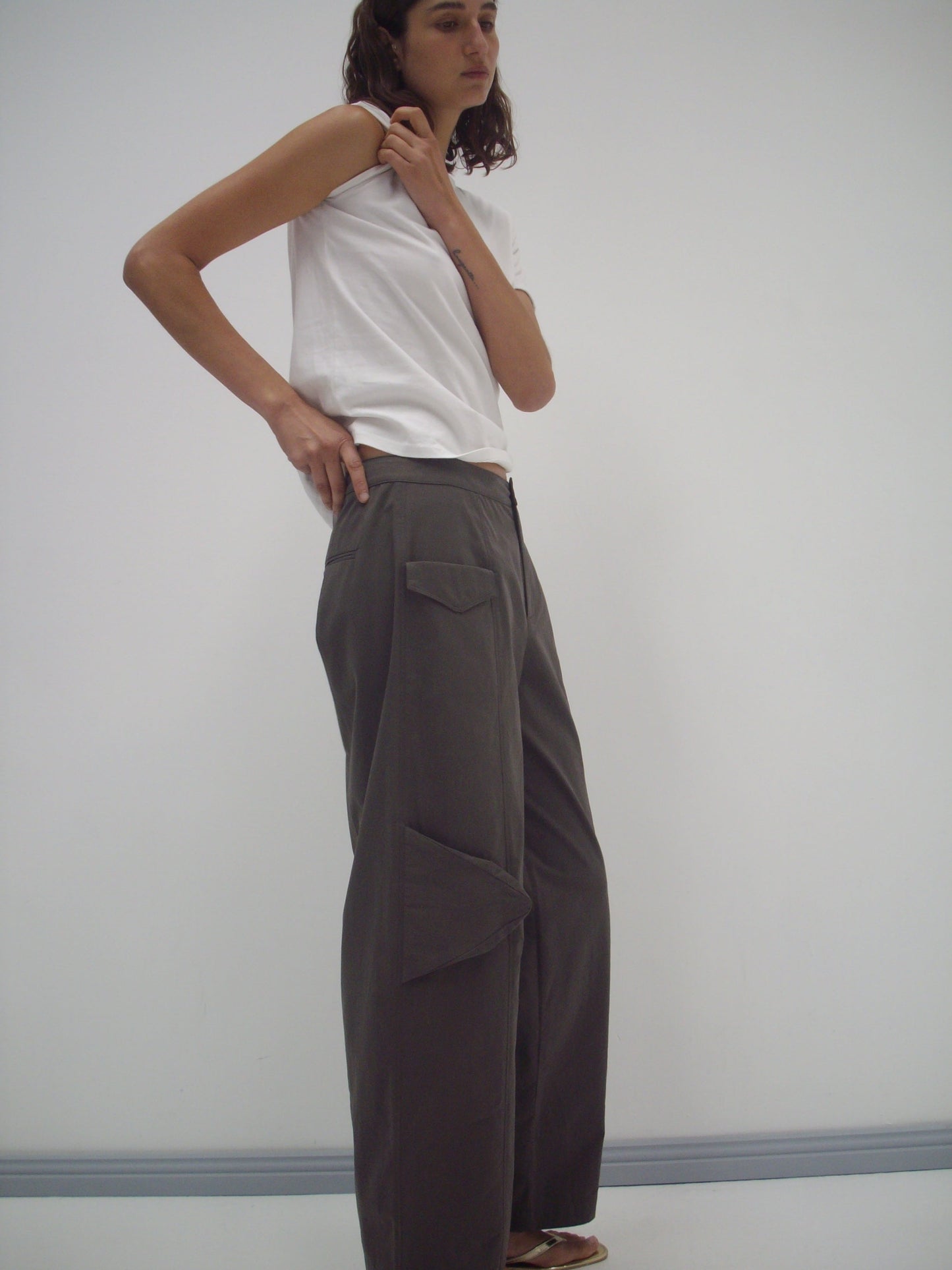 Someone stands against a plain white wall, wearing a sleeveless white top and the Pocket Pant - Wood, which are loose-fitting gray organic cotton twill pants with pockets. Their shoulder-length wavy hair and hand on hip add a casual touch.