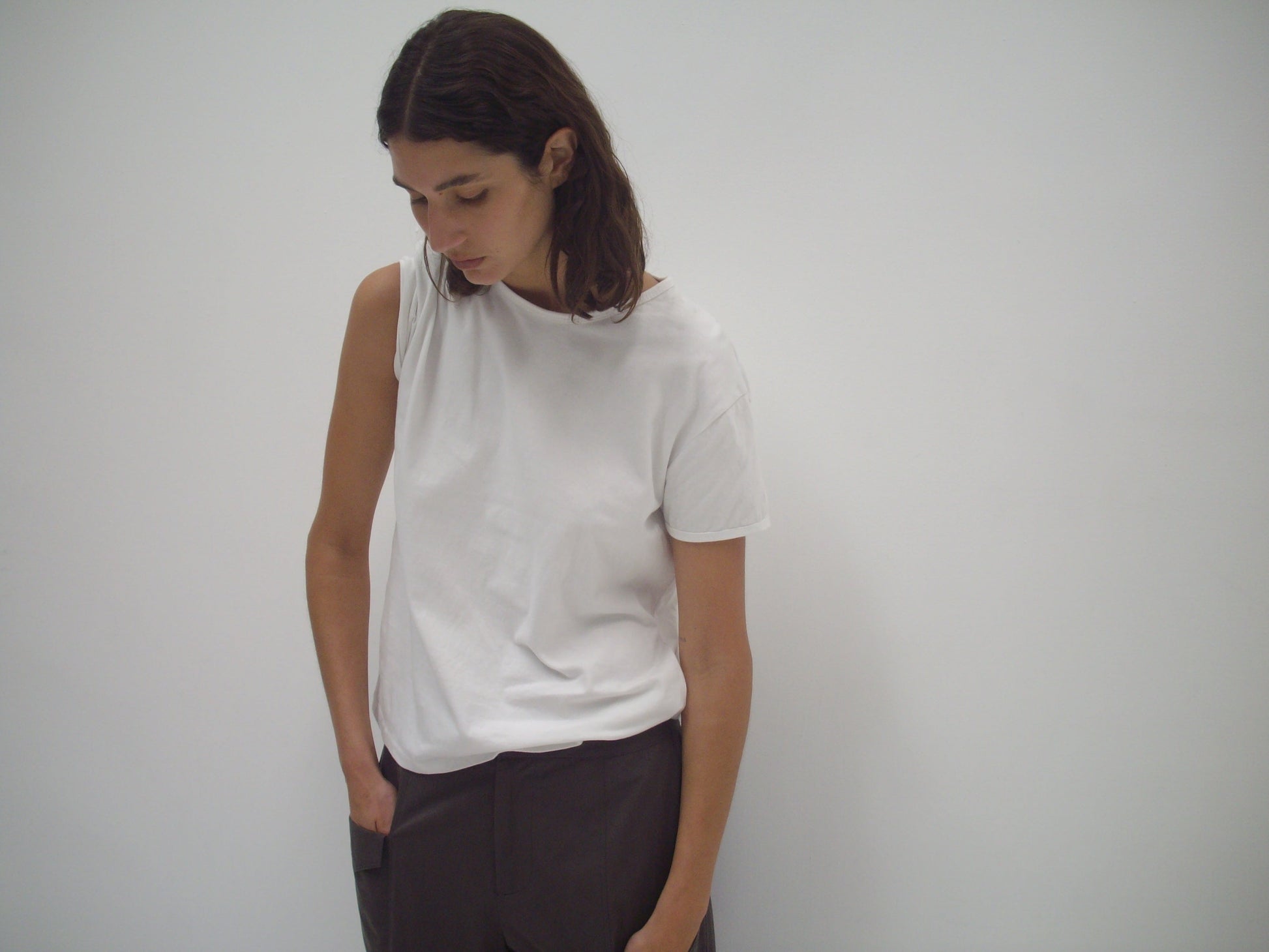 A person with shoulder-length brown hair wears the Simple Tee - White by Deiji Studios and dark pants, standing against a blank white wall and looking down with one hand in their pocket.