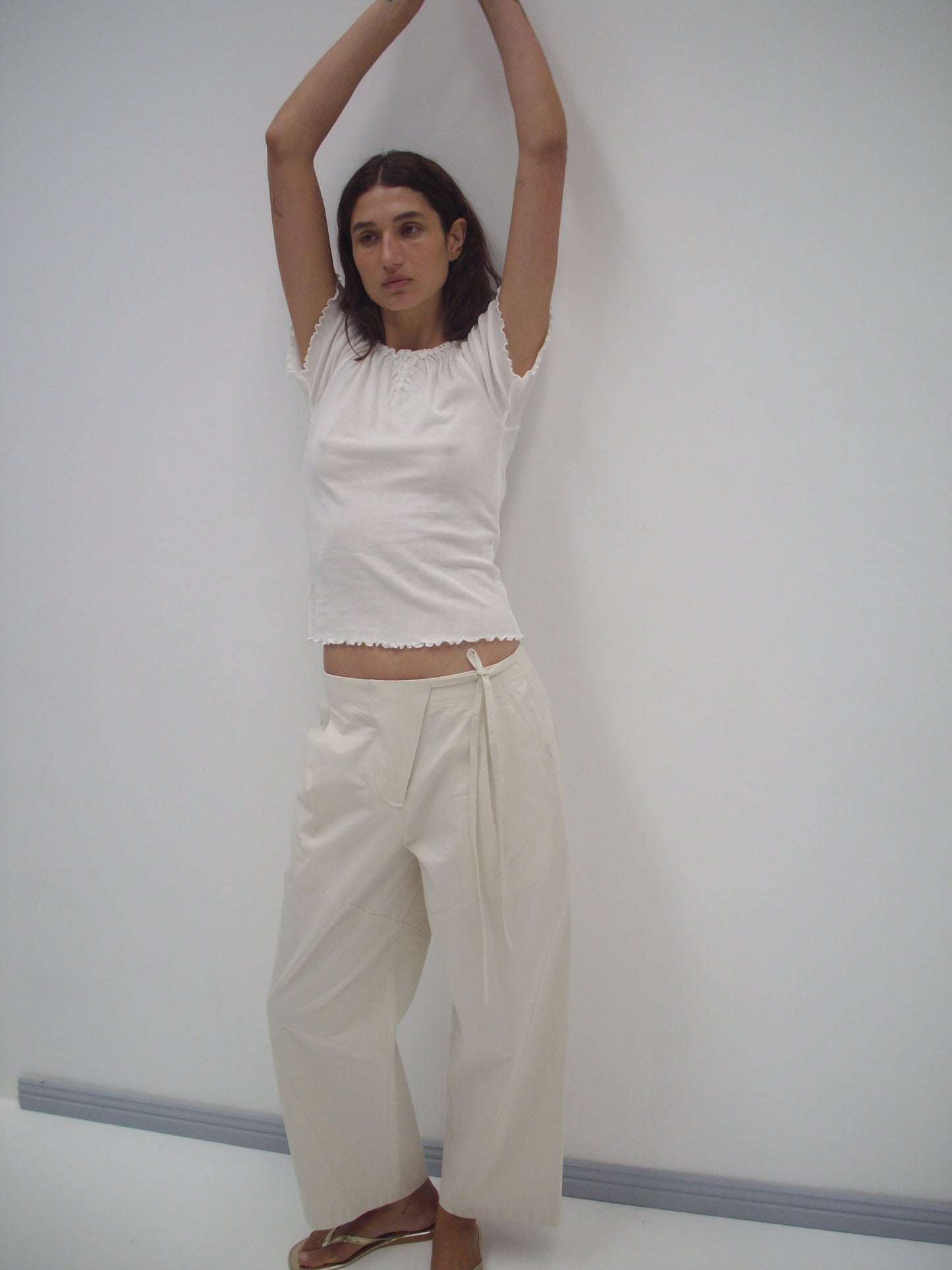 A person with long dark hair wears a white short-sleeve top, the Flag Tie Pant in Off White with a loose fit, and sandals, standing against a plain white wall with arms raised above their head.