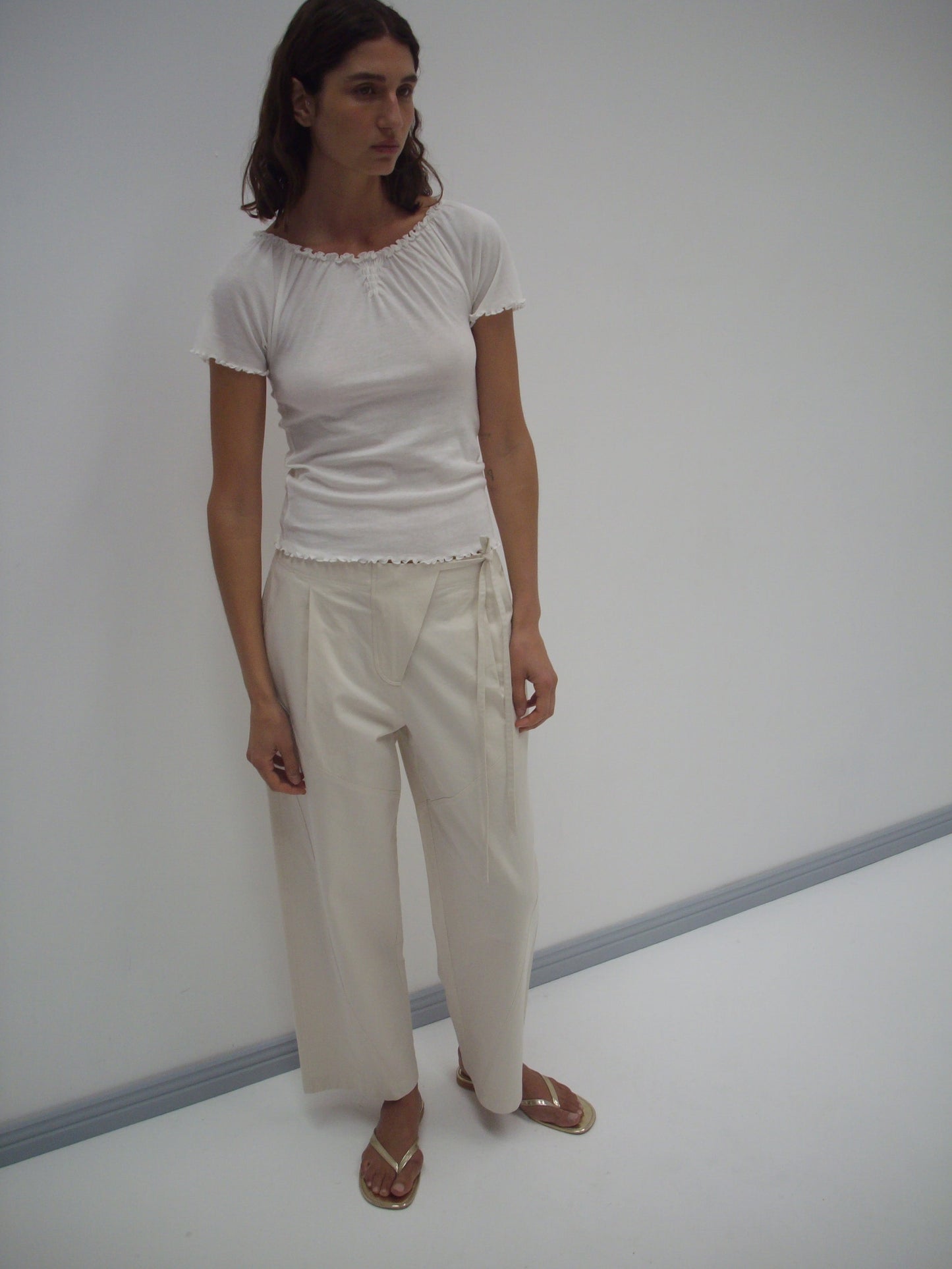 A person stands against a plain white wall, wearing a fitted short-sleeve white top, the Flag Tie Pant in Off White, and sandals. Their expression is neutral and their arms hang loosely at their sides.