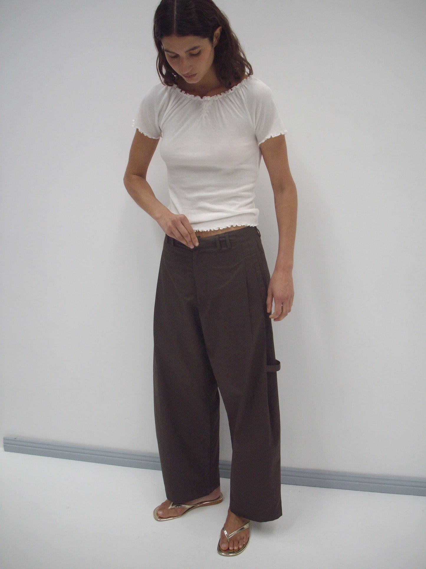 A woman stands against a white wall wearing a fitted white short-sleeve top, the Tab Pant in Dark Brown by Deiji Studios, and gold sandals. She looks down, adjusting the waistband of her pants with one hand.