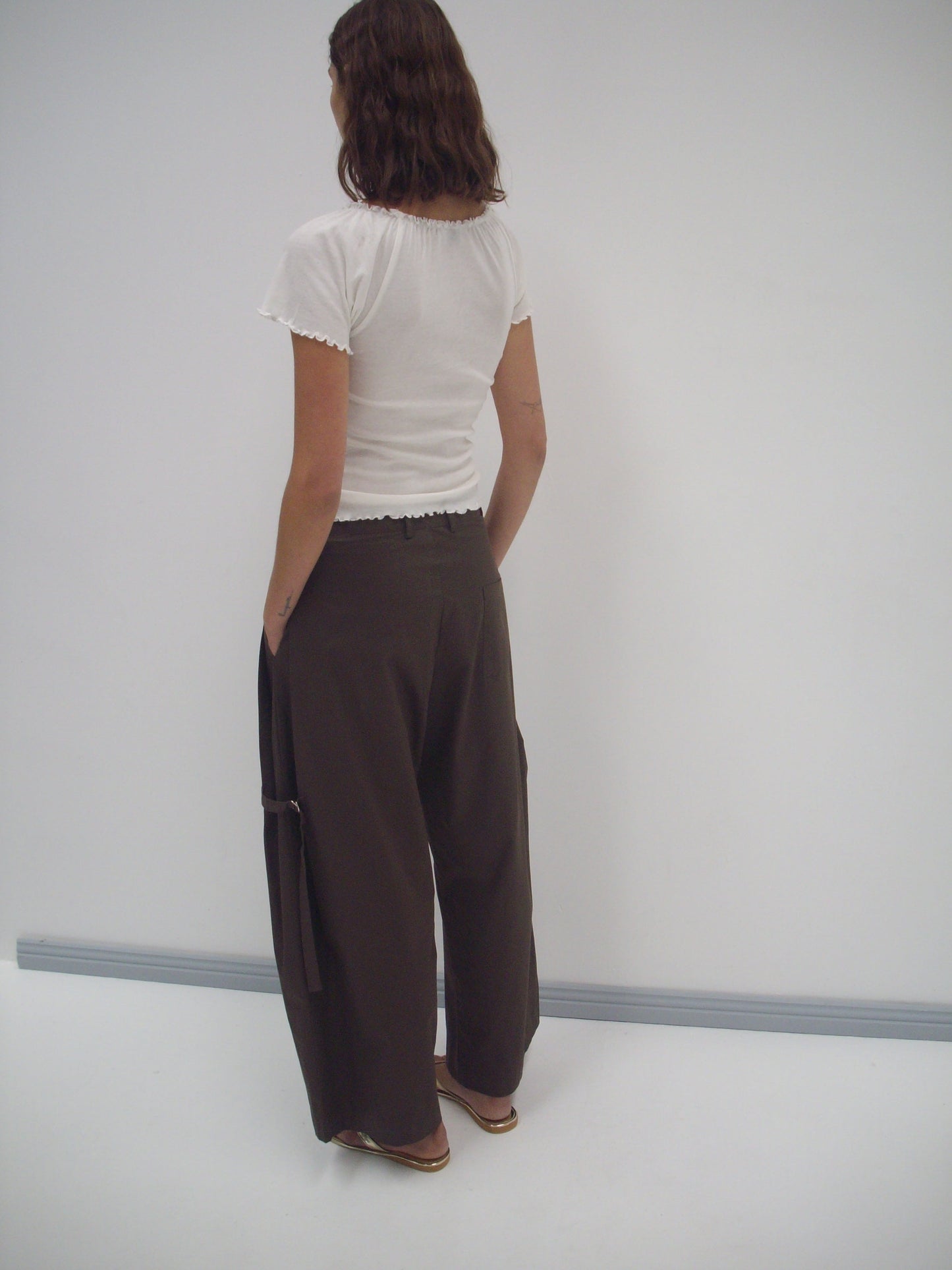 A person with wavy brown hair stands facing away, wearing a short-sleeved white top, the Tab Pant in Dark Brown from Deiji Studios with side straps, and sandals, set against a plain white wall and floor.
