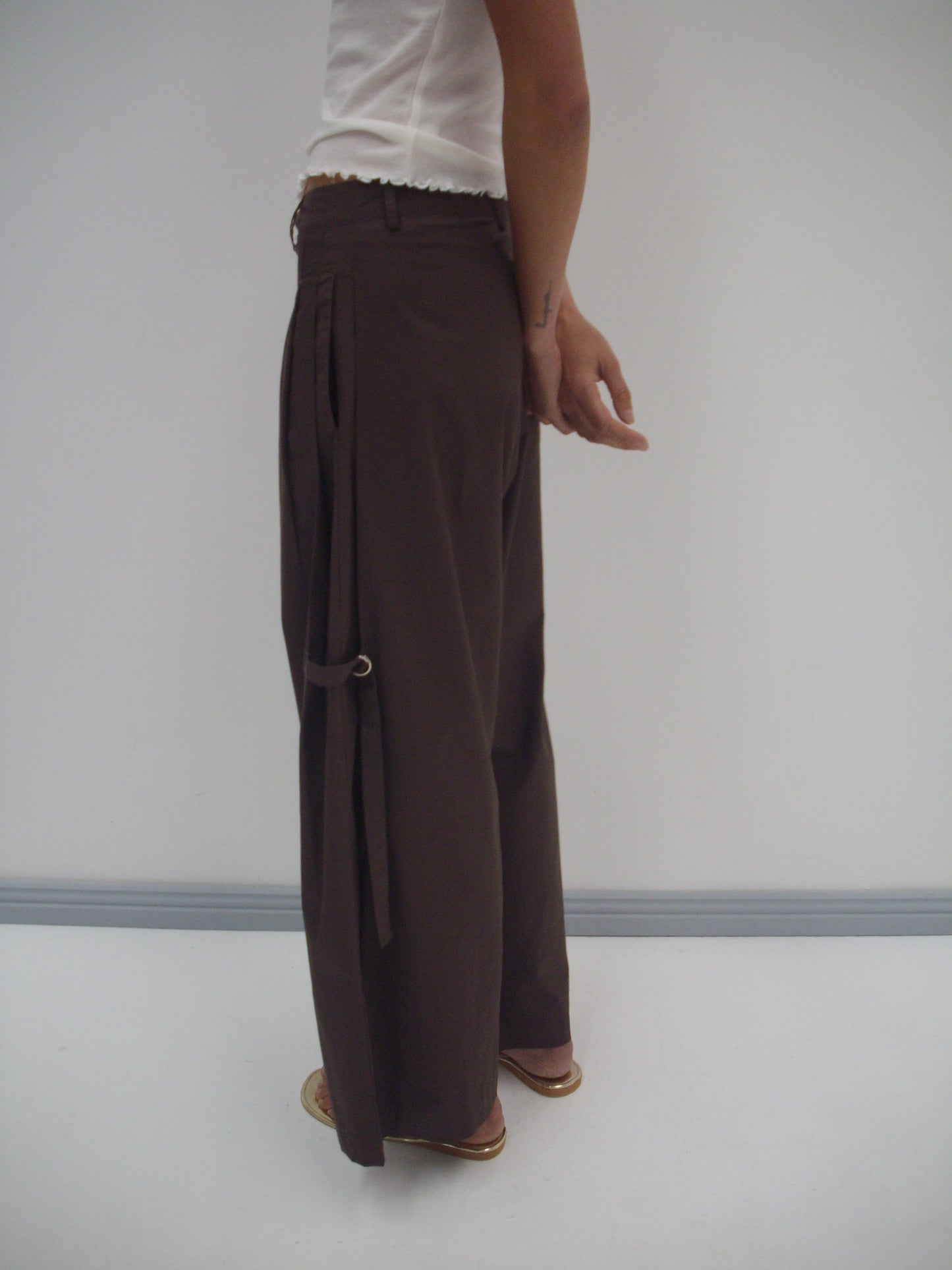 Wearing the Deiji Studios Tab Pant in dark brown organic cotton with side straps, styled with a white top. The photo shows only the lower half of the body, standing sideways against a plain white wall.