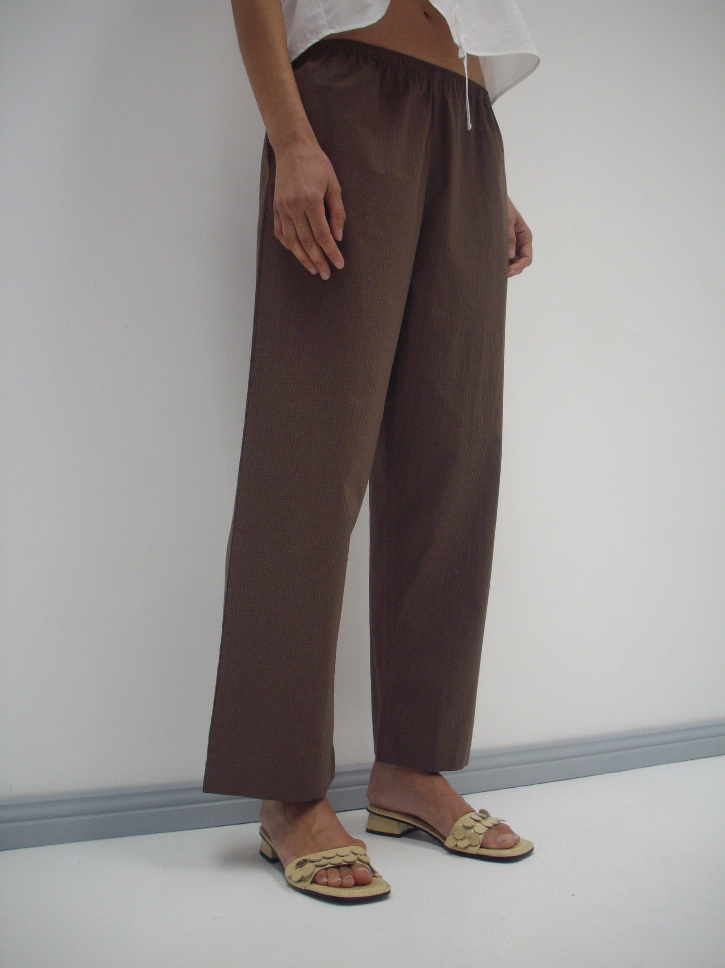 A person wearing the Ease Trouser - Carob Check, featuring a loose fit and elastic waist, pairs them with a white top and light open-toe sandals while standing against a plain white wall. Photo cropped at chest.