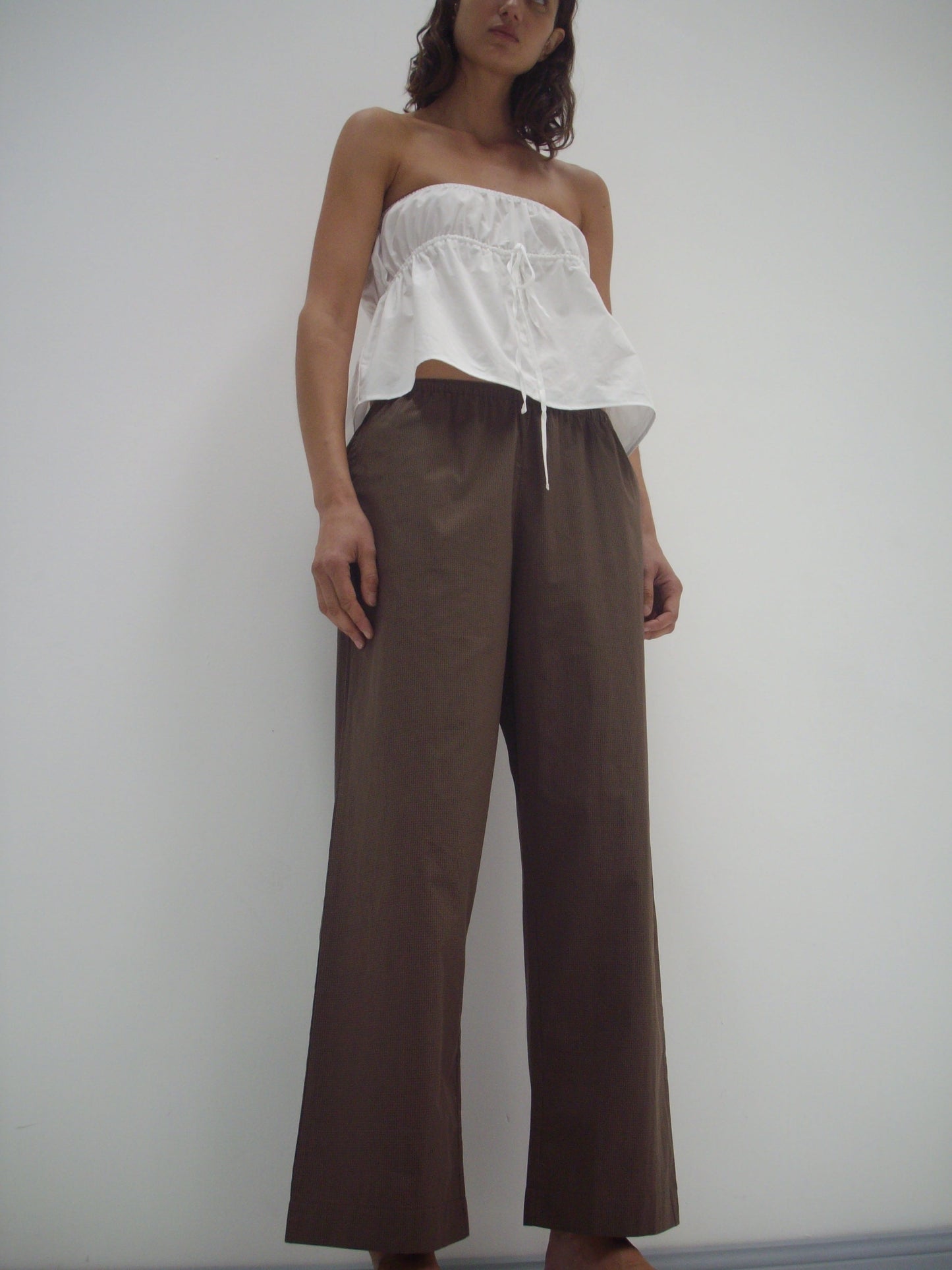 A person stands against a white wall, wearing a strapless top and the Ease Trouser - Carob Check. The low-angle photo captures the effortless style and comfort of these elastic waist, wide-legged trousers.