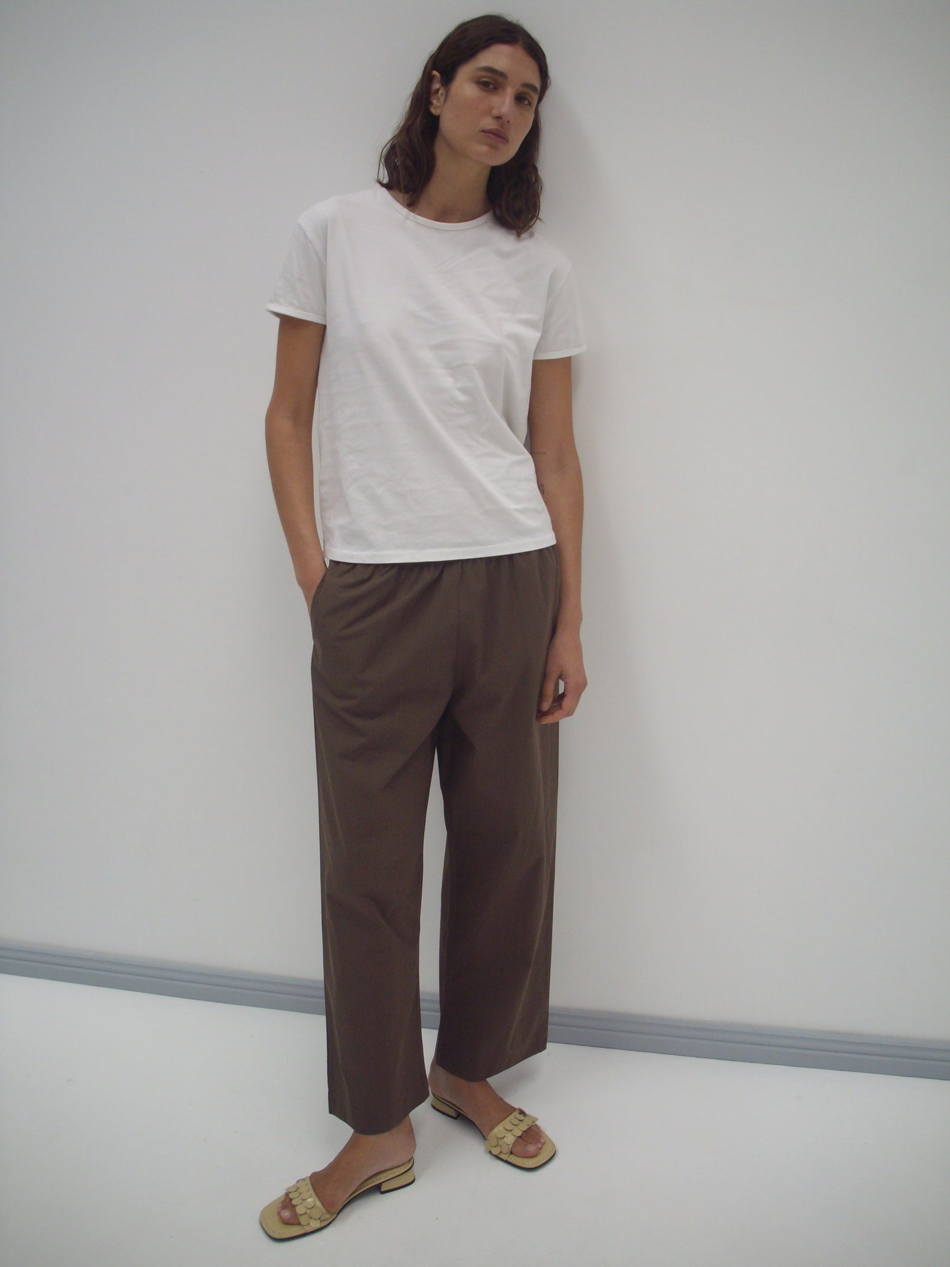 A person with long dark hair stands against a white wall, wearing the Simple Tee - White from Deiji Studios, loose brown trousers, and beige sandals. Their hands are in their pockets and they have a neutral expression.