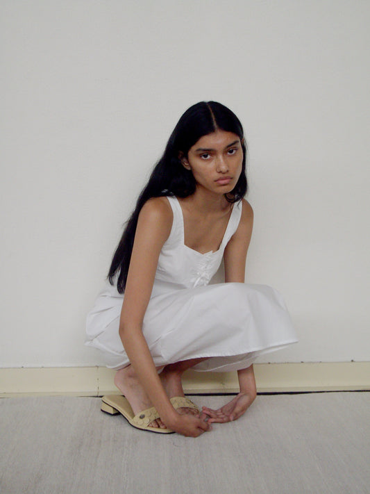 A woman with long dark hair, wearing the Eyelet Dress, crouches on the floor against a plain white wall and looks into the camera with a neutral expression.