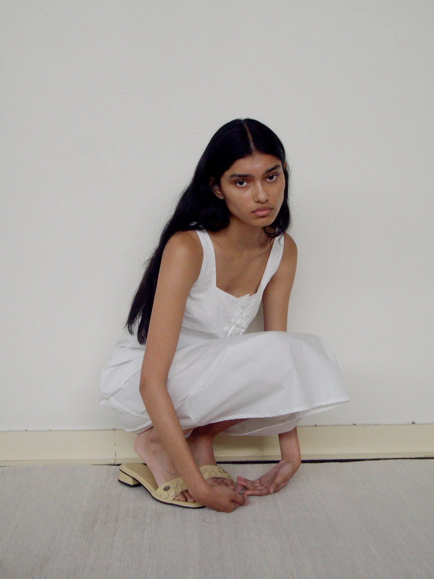 A young woman with long dark hair wears the Eyelet Dress, squatting on the floor and gazing thoughtfully at the camera against a plain white wall.