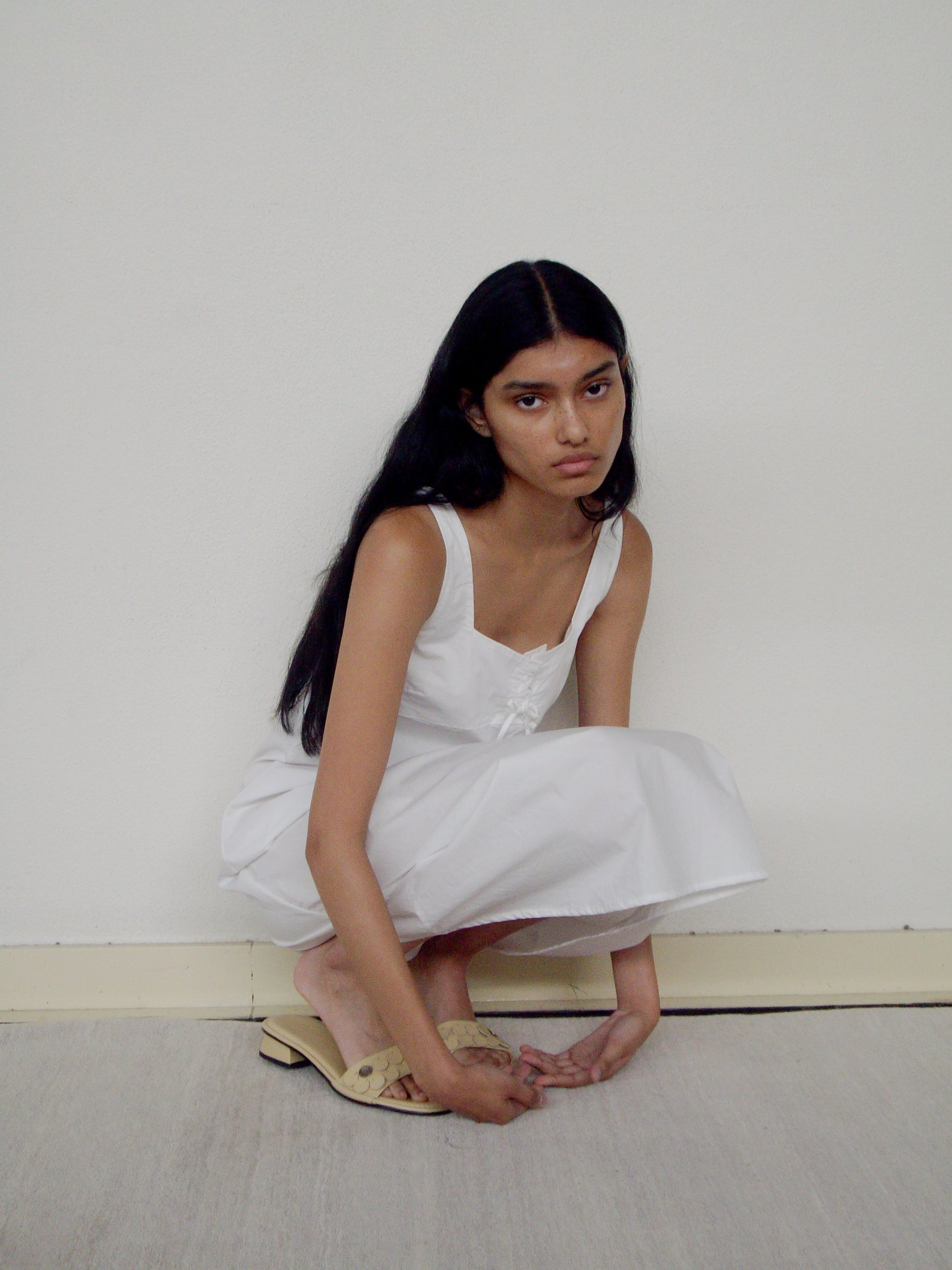 A young woman with long dark hair wears the Eyelet Dress, squatting on the floor and gazing thoughtfully at the camera against a plain white wall.