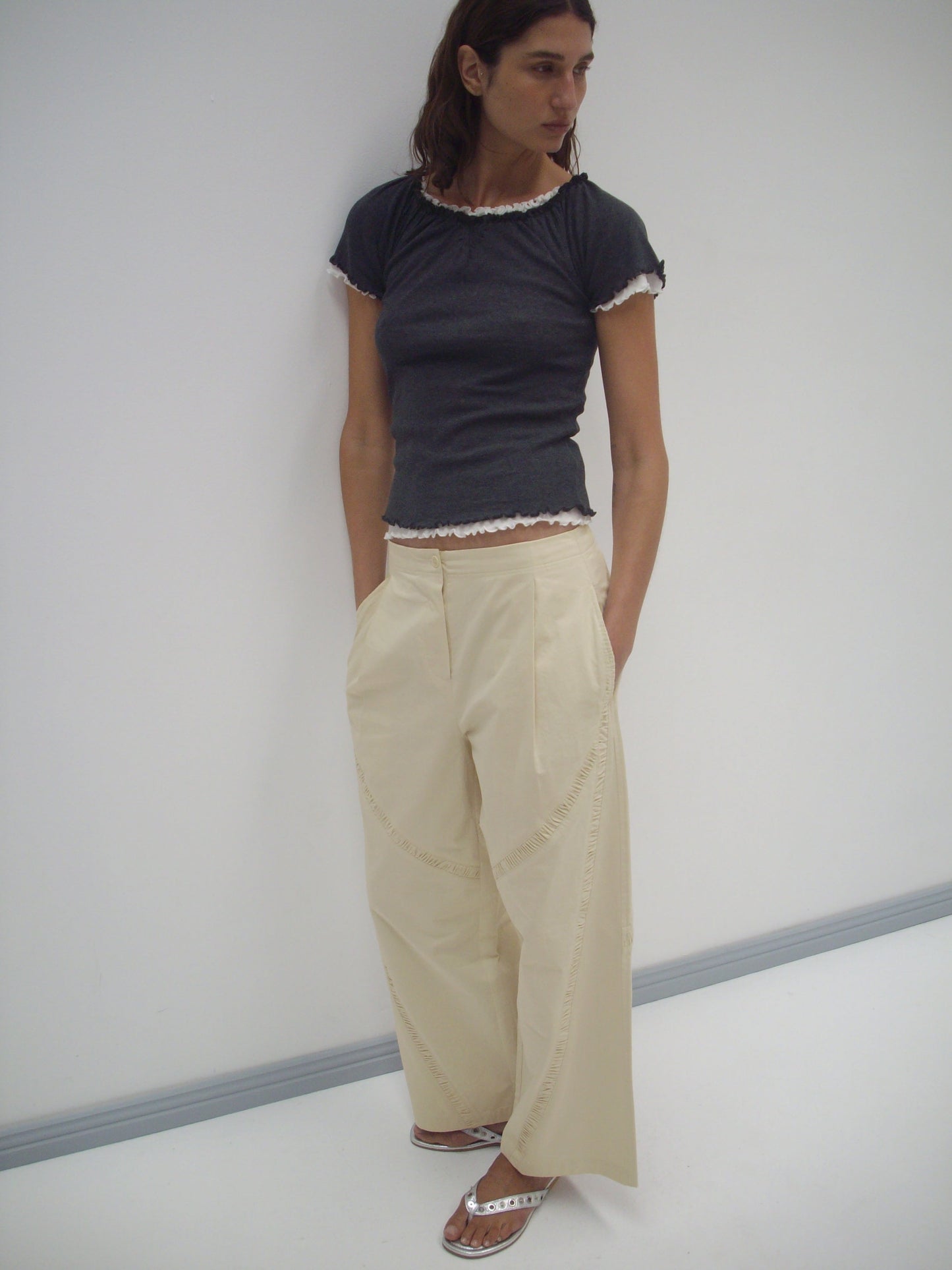 A person stands against a white wall wearing a fitted dark gray t-shirt with white trim, Deiji Studios’ Channel Pant in Stone (relaxed, straight-leg organic cotton twill), and white sandals, striking a relaxed pose with one hand in their pocket.