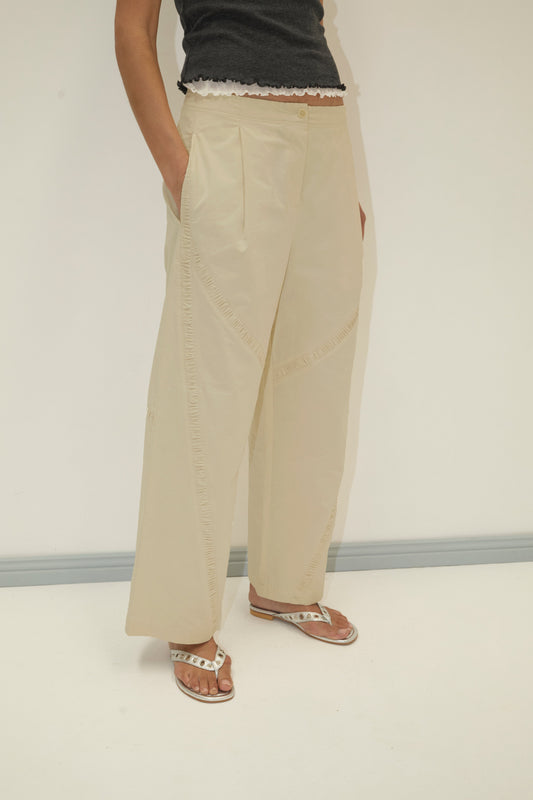 A person stands against a plain background wearing the Channel Pant - Stone in organic cotton twill with relaxed cocoon legs, paired with a black scallop-hem top and white sandals. Their hands are in their pockets; their head is out of frame.