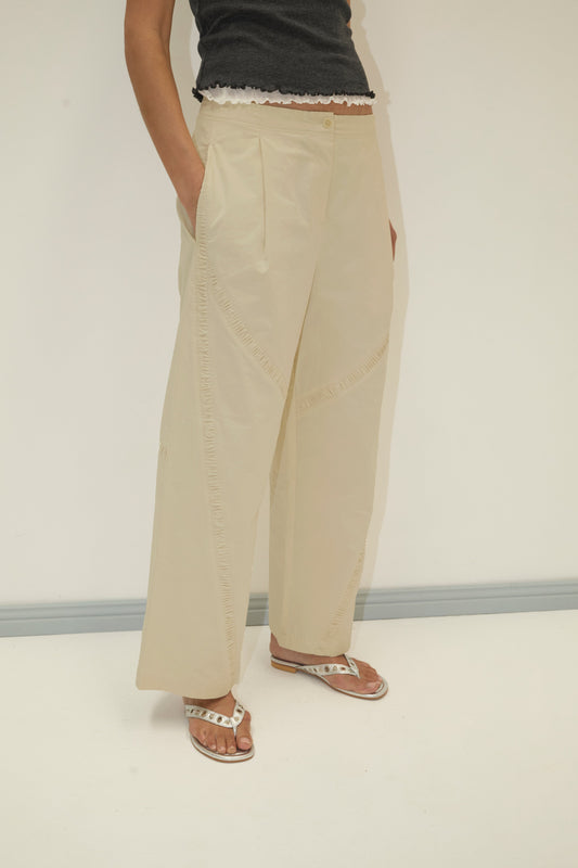 Wearing the Channel Pant - Stone by Deiji Studios, relaxed straight leg in organic cotton twill, paired with a black ruffled hem top and white sandals, a person stands against a plain white background with hands in pockets.