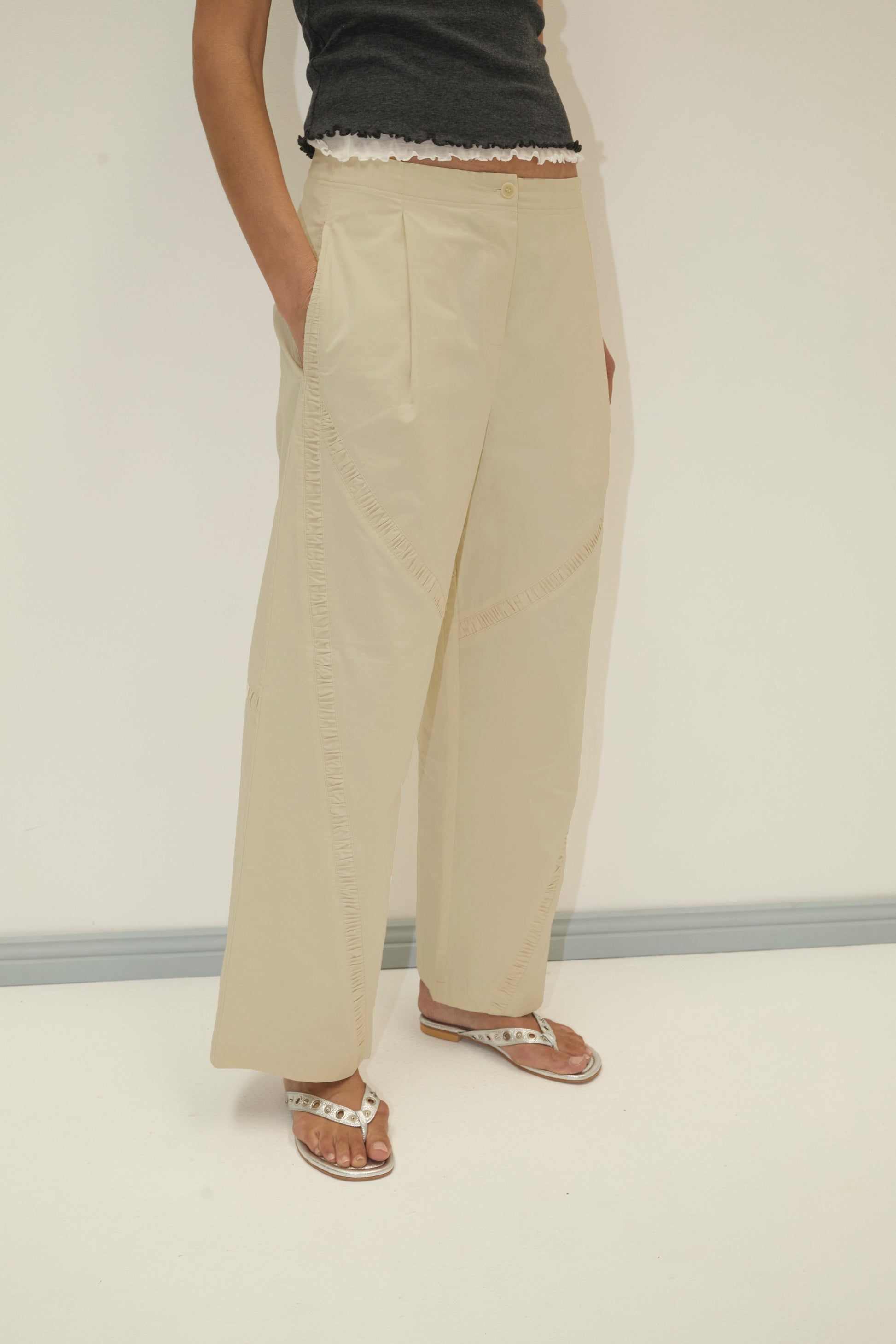 A person stands against a plain background wearing the Channel Pant - Stone in organic cotton twill with relaxed cocoon legs, paired with a black scallop-hem top and white sandals. Their hands are in their pockets; their head is out of frame.