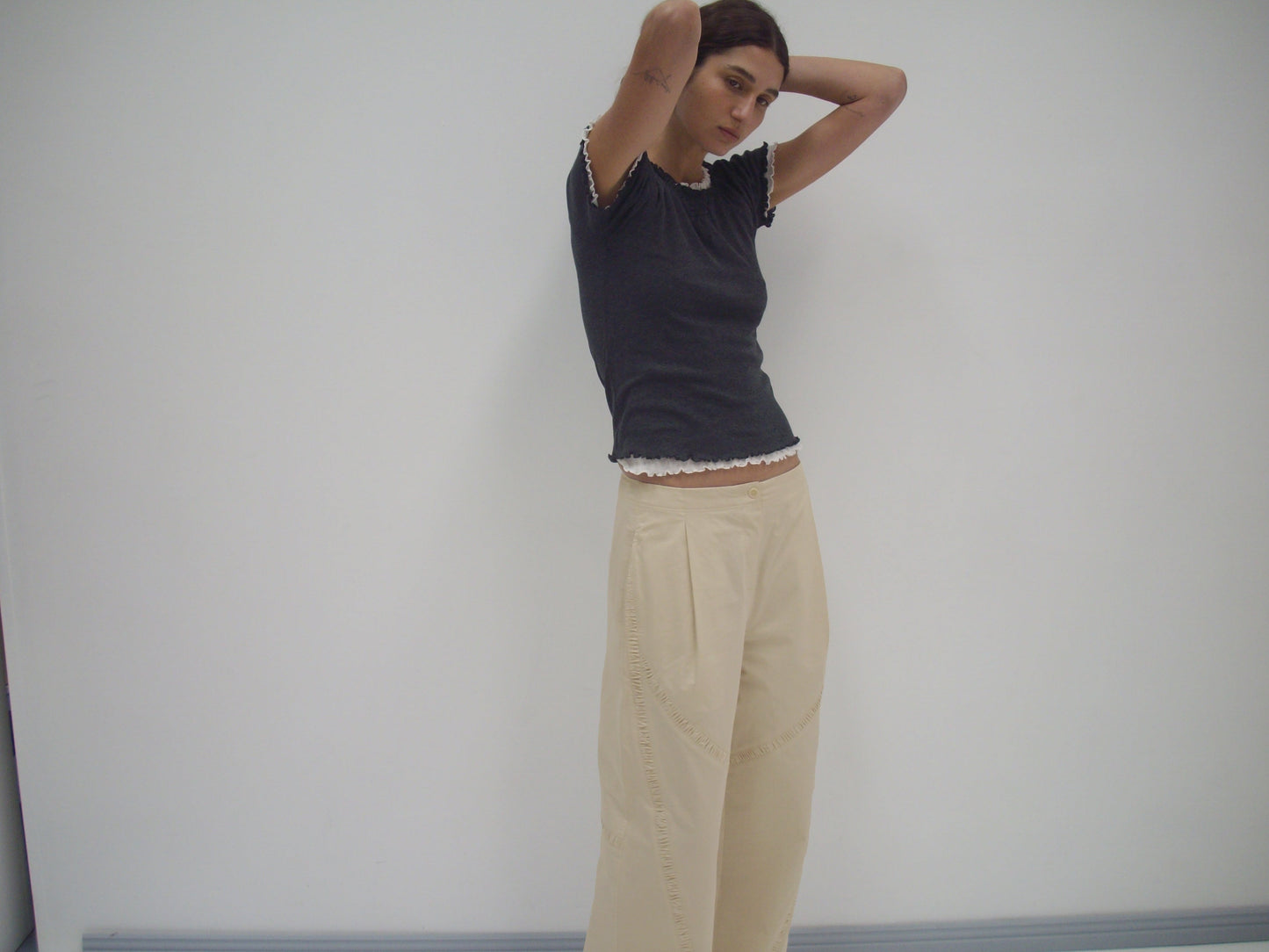 A person stands against a plain white wall with hands behind their head, wearing a black short-sleeve top with white trim and the Channel Pant - Stone in organic cotton twill from Deiji Studios.