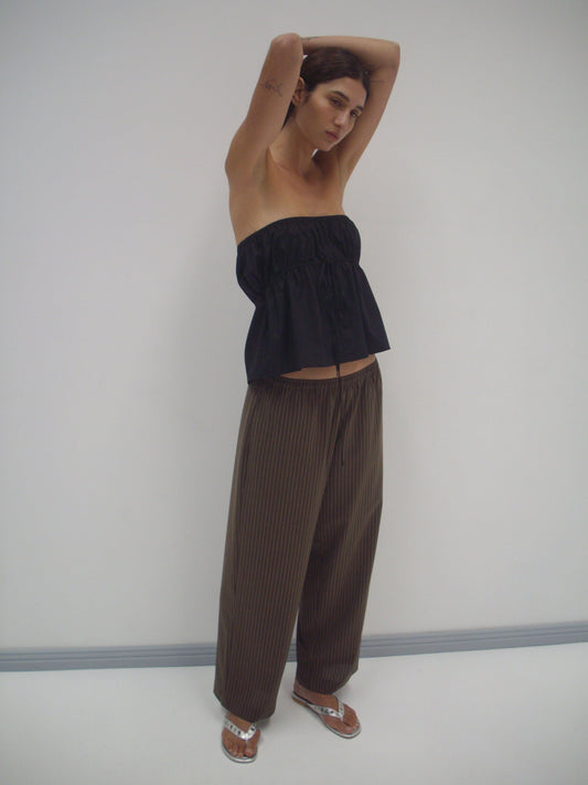 A person stands against a white wall, wearing Deiji Studios’ black strapless cotton top, loose brown pinstriped pants, and white sandals, with arms raised above their head.