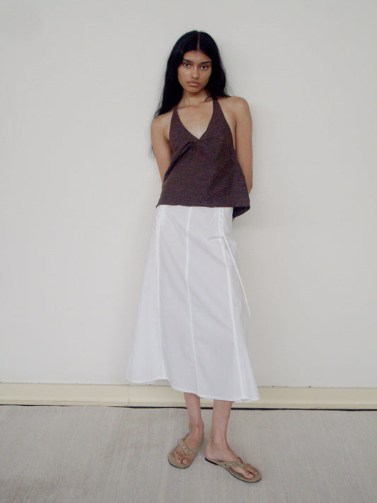 A woman with long dark hair stands against a white wall, wearing a brown halter top, tan sandals, and the Double Lace Up Skirt—a white midi skirt with side ties. Her arms are behind her back as she looks directly at the camera.