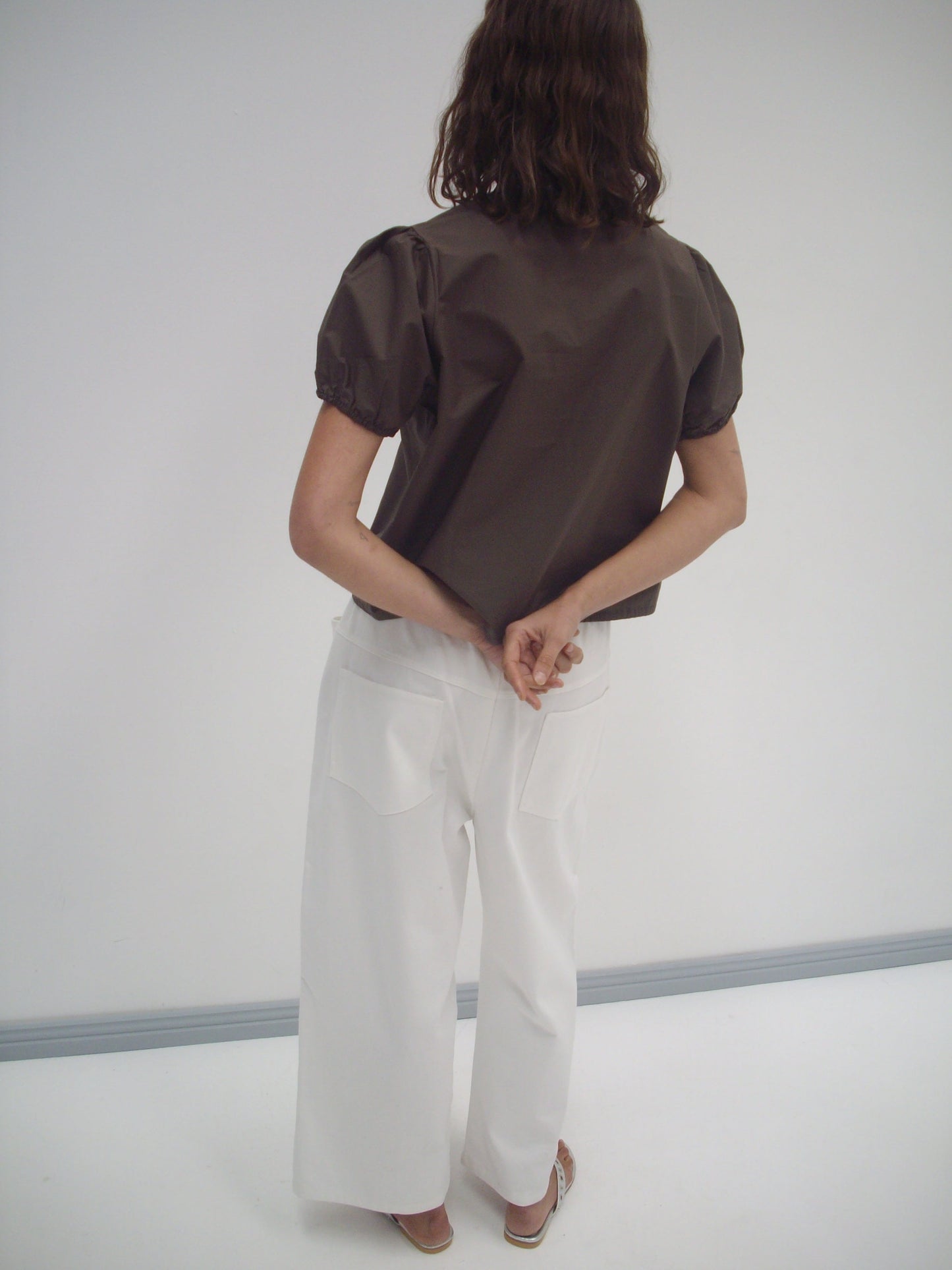 A person with wavy brown hair, wearing a brown short-sleeve top and the Square Pocket Pant in white, stands facing away with hands clasped behind their back against a plain white background.