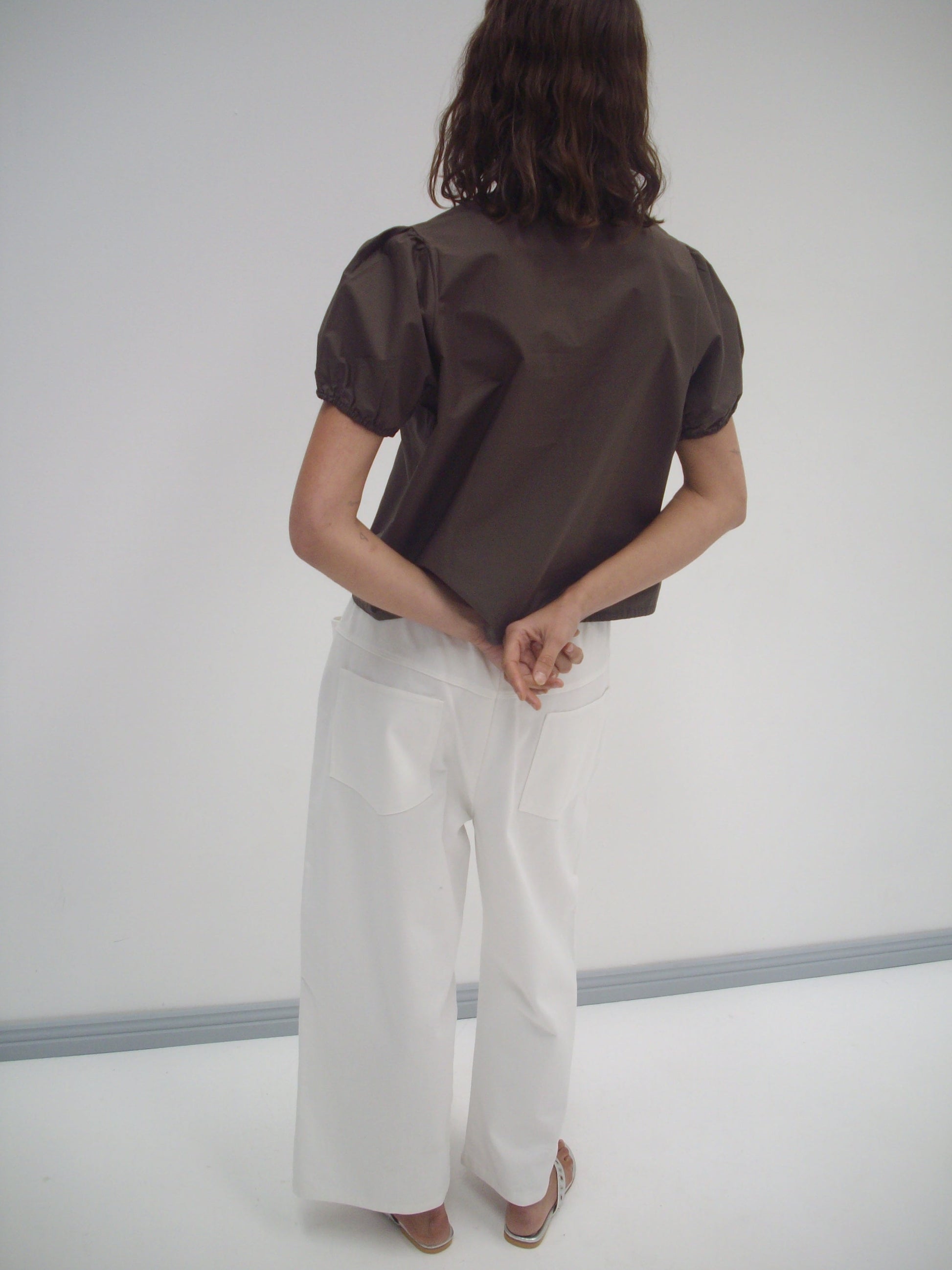 A person with wavy brown hair, wearing a brown short-sleeve top and the Square Pocket Pant in white, stands facing away with hands clasped behind their back against a plain white background.