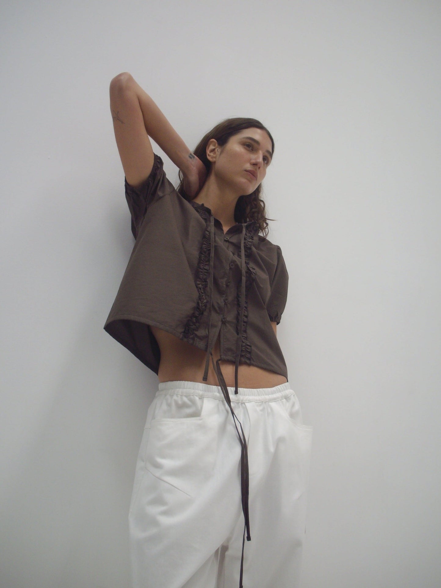 A person stands against a plain white wall, wearing the Double Frill Top - Chocolate from Deiji Studios and white pants. One arm is raised behind their head, and they appear relaxed and confident.