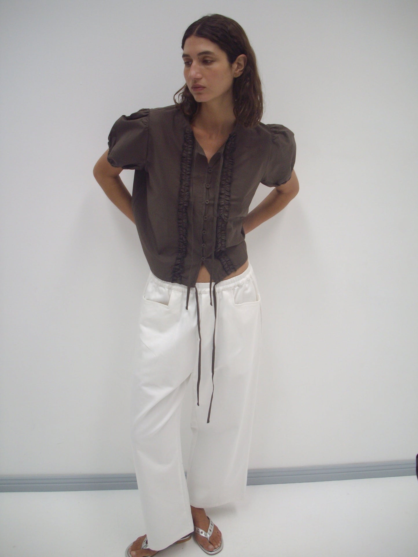 A woman with medium-length dark hair stands against a plain white wall, wearing Deiji Studios’ Double Frill Top in Chocolate with wide-legged white pants, looking to the side and resting her hands in her pockets.