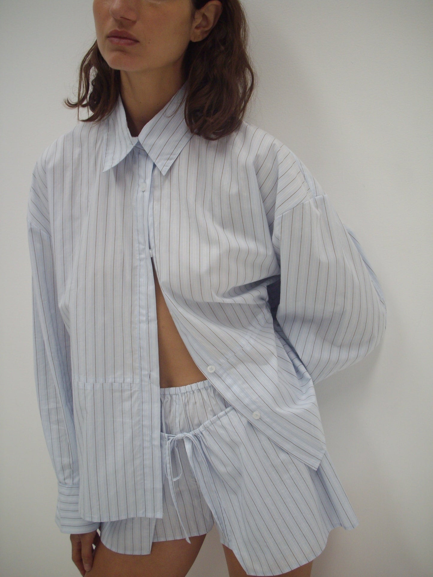 A woman stands against a plain white background wearing the Deiji Studios Room Shirt - Mineral Stripe, left partially open to reveal matching drawstring shorts underneath. Her face is partially out of frame.