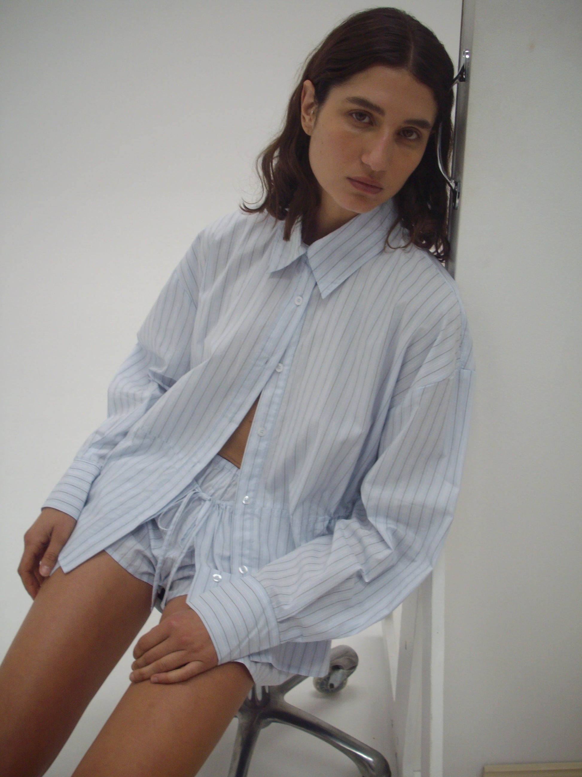 A person with wavy brown hair wears an oversized, light blue striped button-up shirt and the Deiji Studios Skort - Mineral Stripe mini skort, sitting on a metal stool against a plain white background.