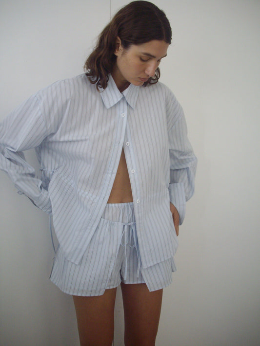 A model wears the Deiji Studios Room Shirt in Mineral Stripe, an oversized long-sleeve pinstripe top, paired with matching drawstring shorts, posed against a white background.