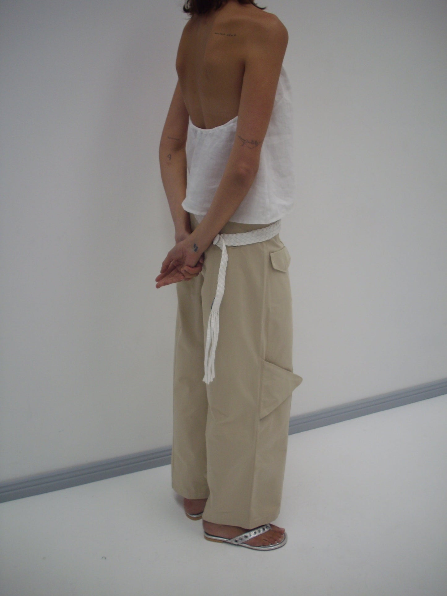 A person stands with their back slightly turned, wearing a white strapless top, Pocket Pant - Stone from Deiji Studios in organic cotton twill, a braided white belt, and white sandals against a plain light background.