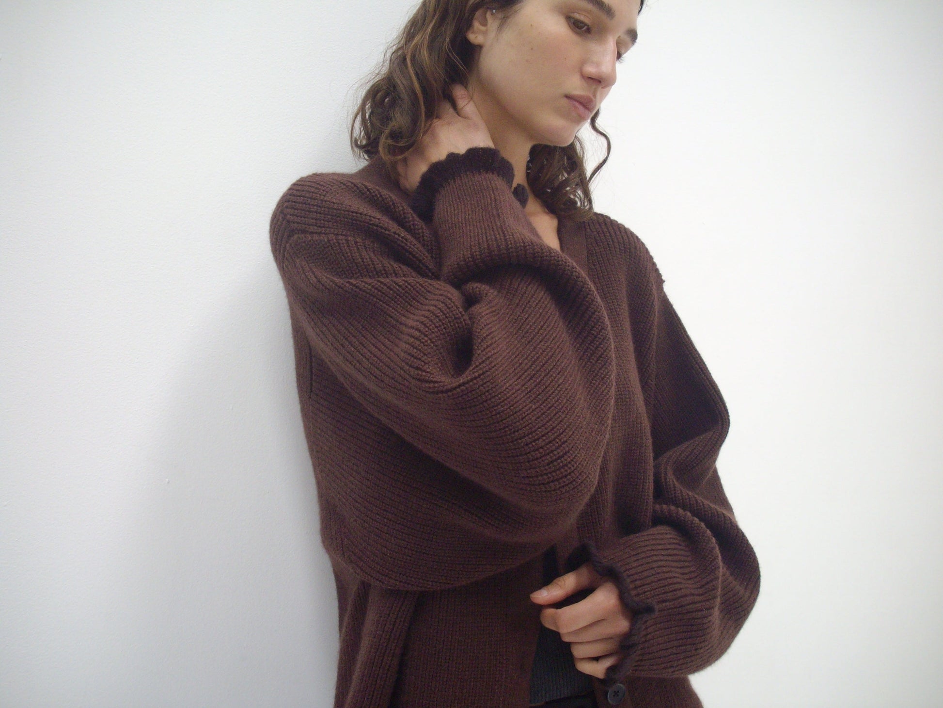 A person with wavy hair wears the Flounce Cardigan - Burgundy by Deiji Studios, made from recycled acrylic, standing against a white wall and looking down thoughtfully with one hand on their neck.