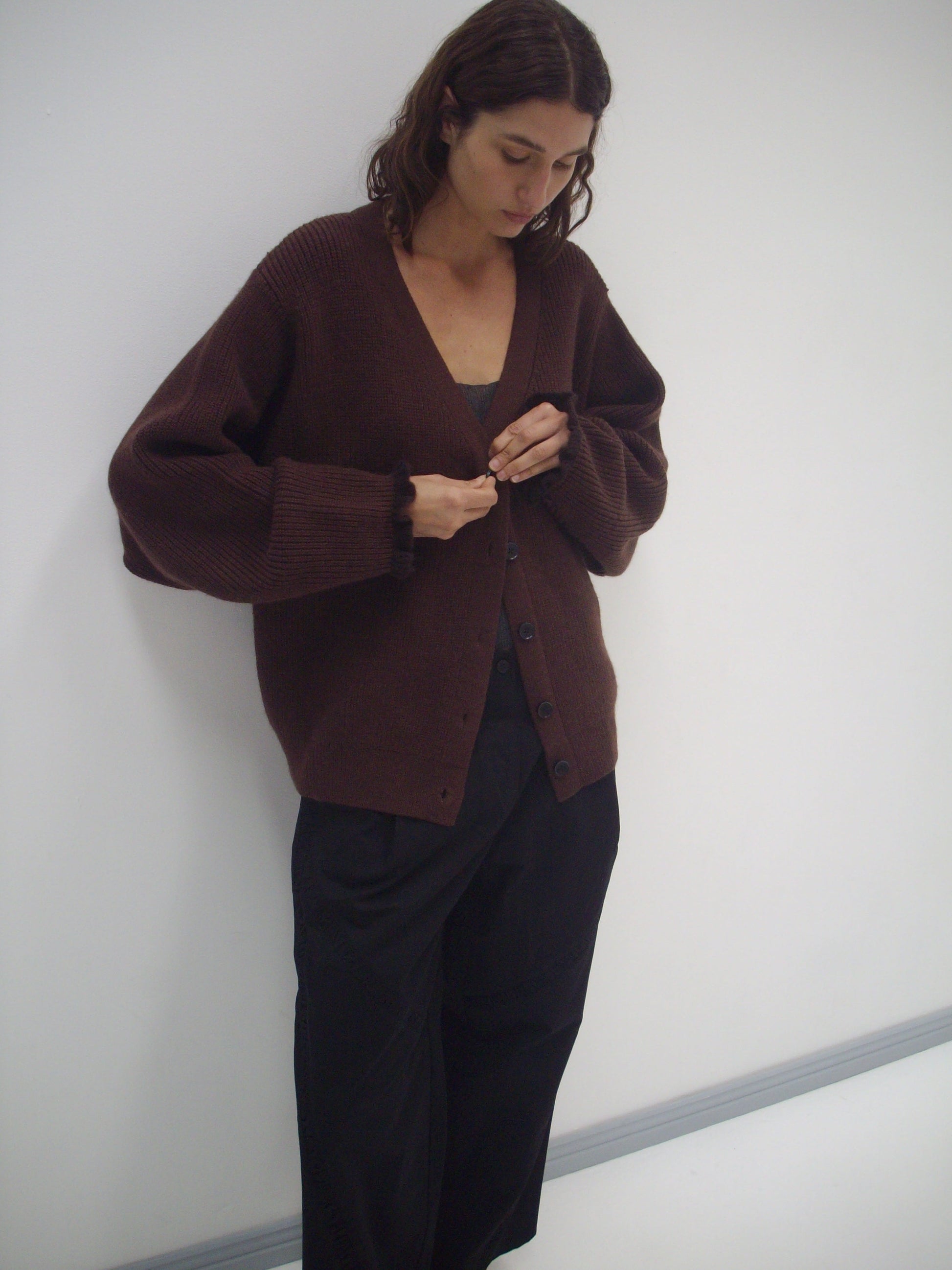 A person with shoulder-length brown hair stands against a white wall, wearing the Flounce Cardigan - Burgundy over a black top and wide-leg black pants, looking down while buttoning the cardigan.
