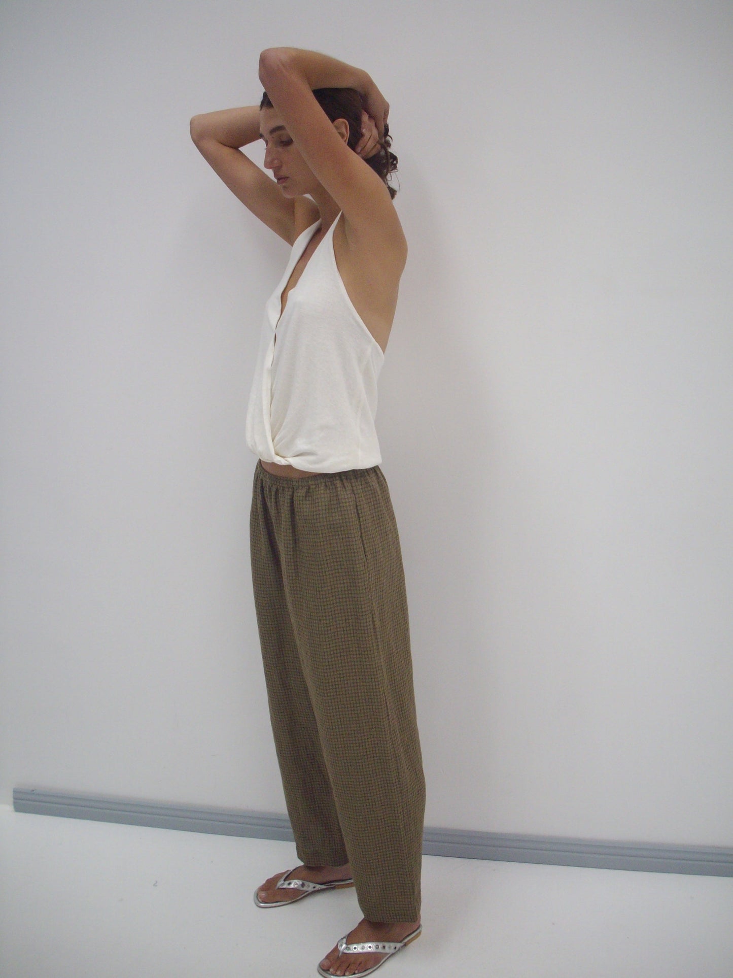 A person stands in a minimalist room, wearing a white sleeveless top, the Linen Ease Trouser - Pine Check, and flip-flops. They have their arms raised with hands behind their head, looking relaxed.