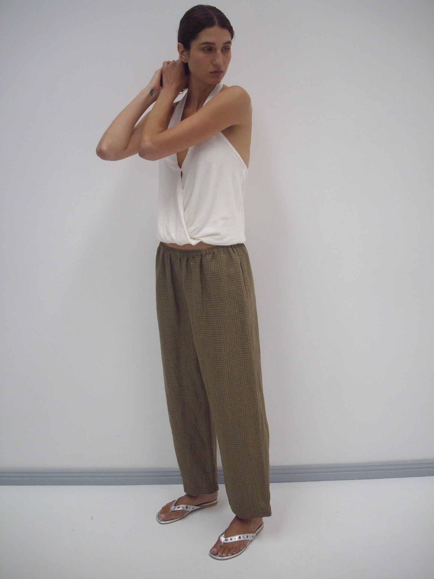 A person stands against a plain white wall, wearing a sleeveless white v-neck top, the Linen Ease Trouser - Pine Check in a loose brown plaid, and silver sandals. They are adjusting their hair with both hands.