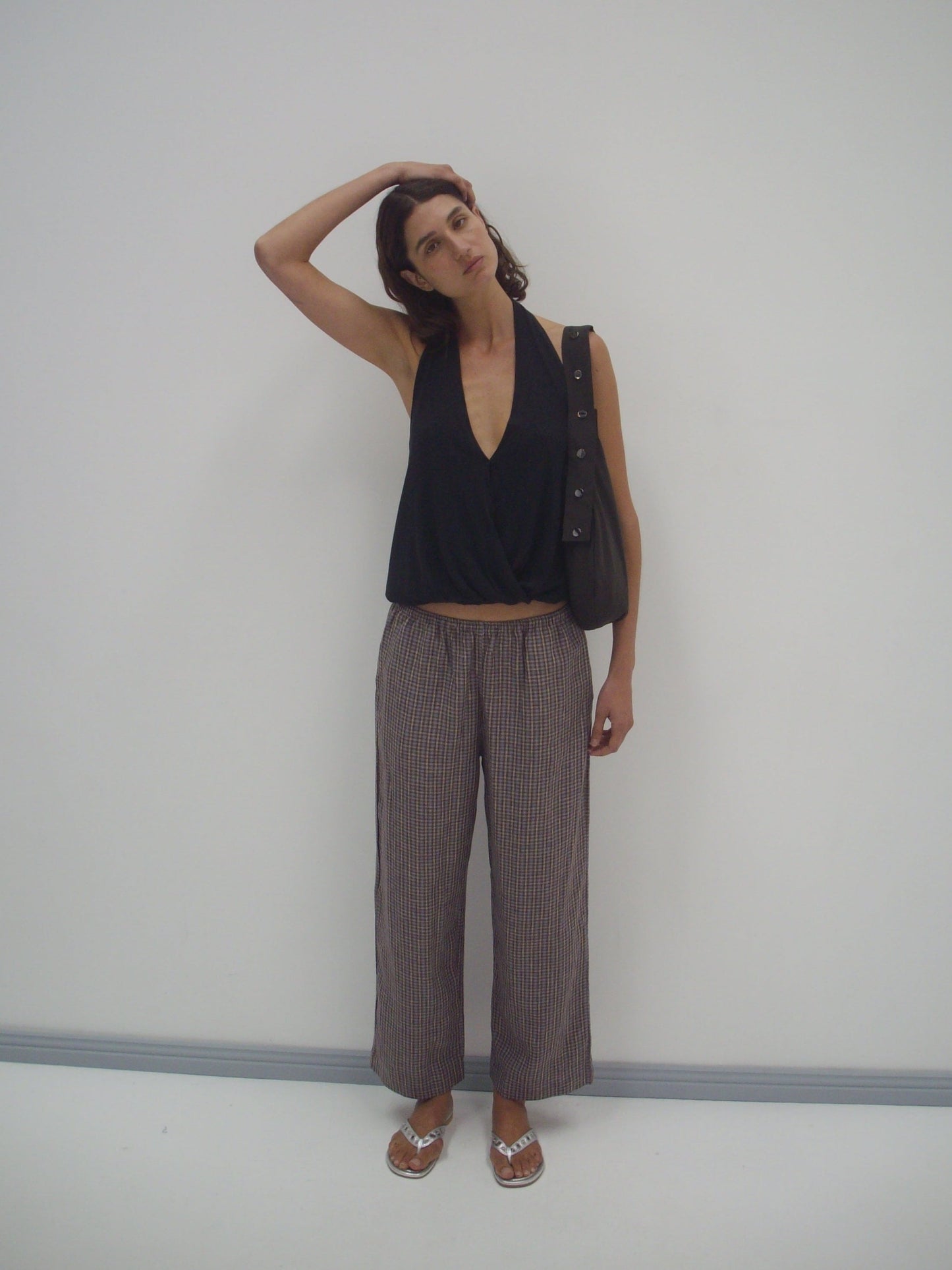 A person stands against a white wall, tilting their head and touching it with one hand. They wear a sleeveless black top, Deiji Studios linen ease trouser in russet check, sandals, and carry a bag over one shoulder.