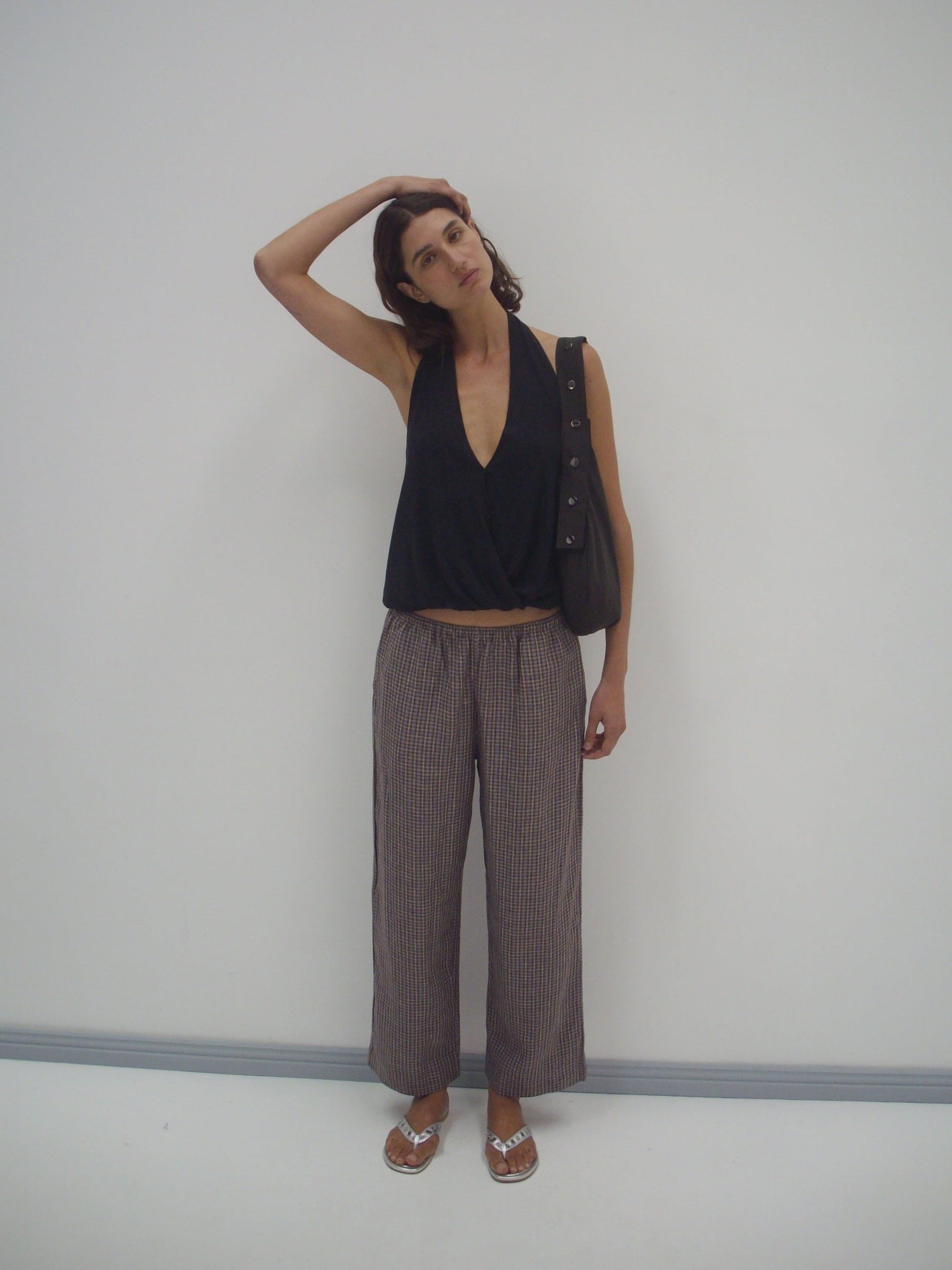 A woman stands against a plain white wall, head tilted with one hand resting on it. She wears the Jersey Top - Black, loose striped pants from Deiji Studios, sandals, and carries a black bag over her shoulder.