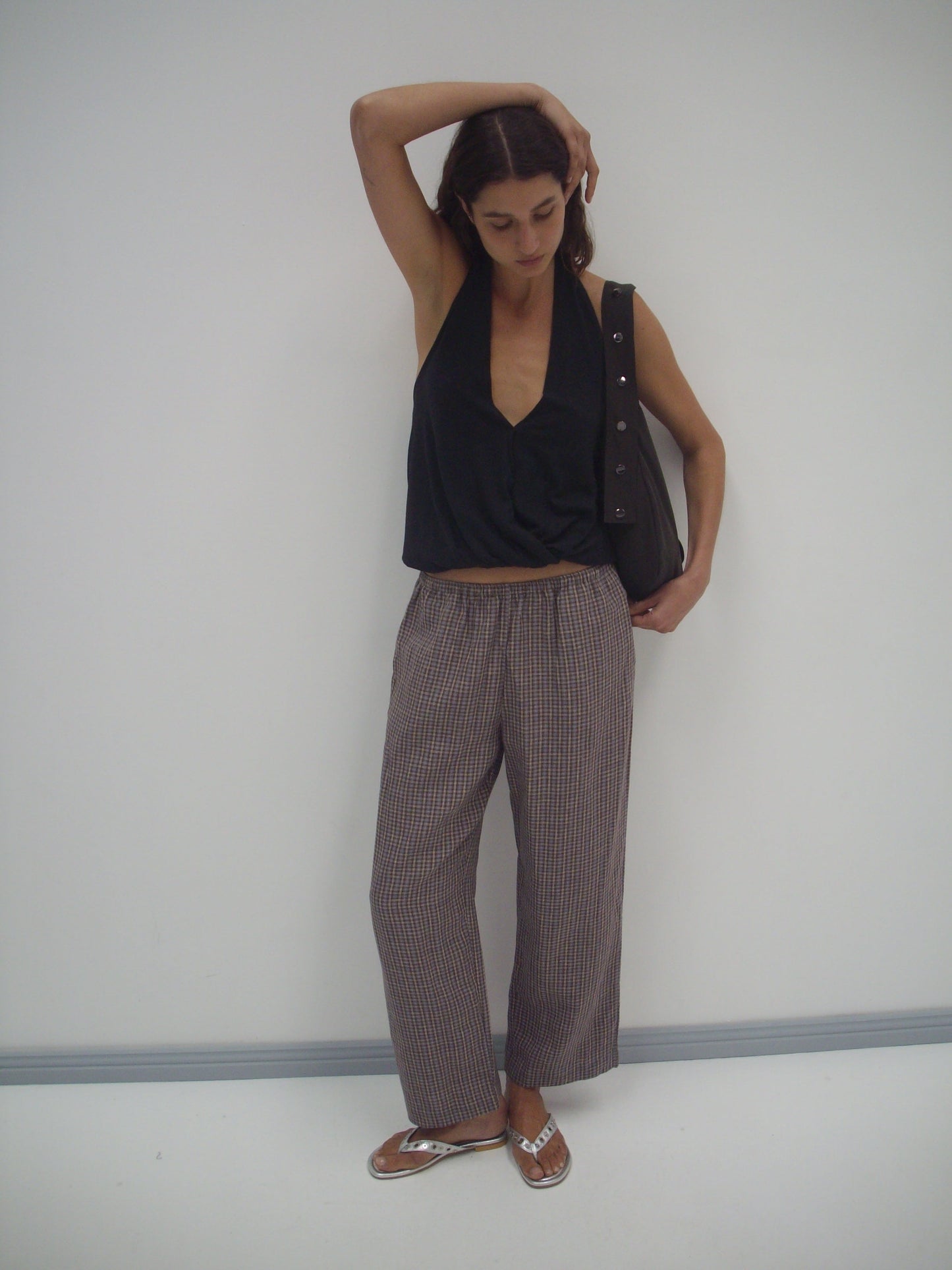 A person stands against a white wall, wearing a sleeveless black top, linen ease trouser - russet check by Deiji Studios, and sandals. They have a bag over one shoulder and touch their head with one arm.